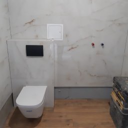 Osoba prywatna - Nowoczesna toaleta wisząca w łazience z marmurowymi płytkami. Widoczna skrzynka narzędziowa, co sugeruje prace wykończeniowe. Podłoga wyłożona panelami.