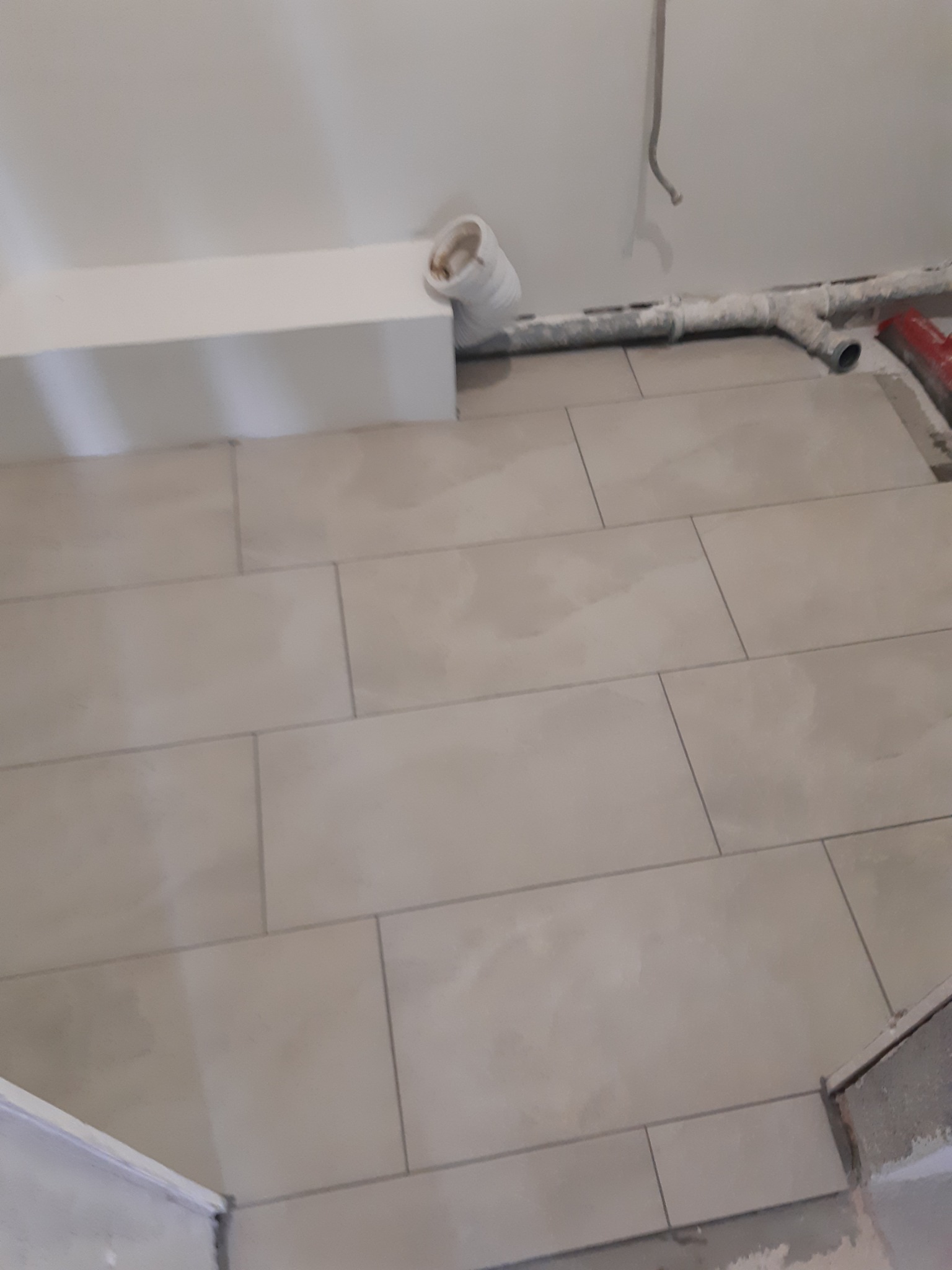 Ułożona, beżowa terakota w łazience, widoczne rury i fragment zabudowy. Świeżo położone płytki podłogowe z fugami, w tle instalacja sanitarna.