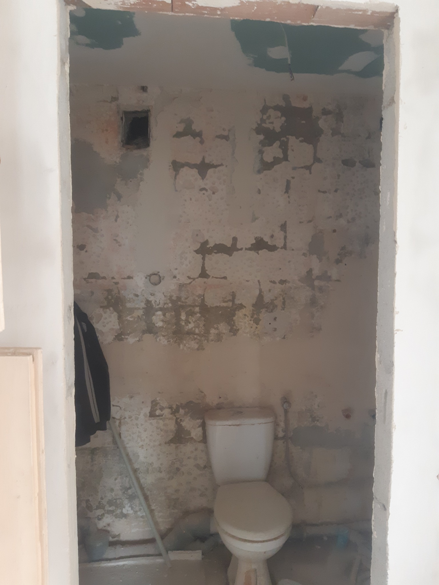 Łazienka w trakcie remontu: surowe ściany z odsłoniętym murem, widoczny stary sedes, otwór wentylacyjny i fragment wiszącej kurtki. Ujęcie z perspektywy wejścia do pomieszczenia.