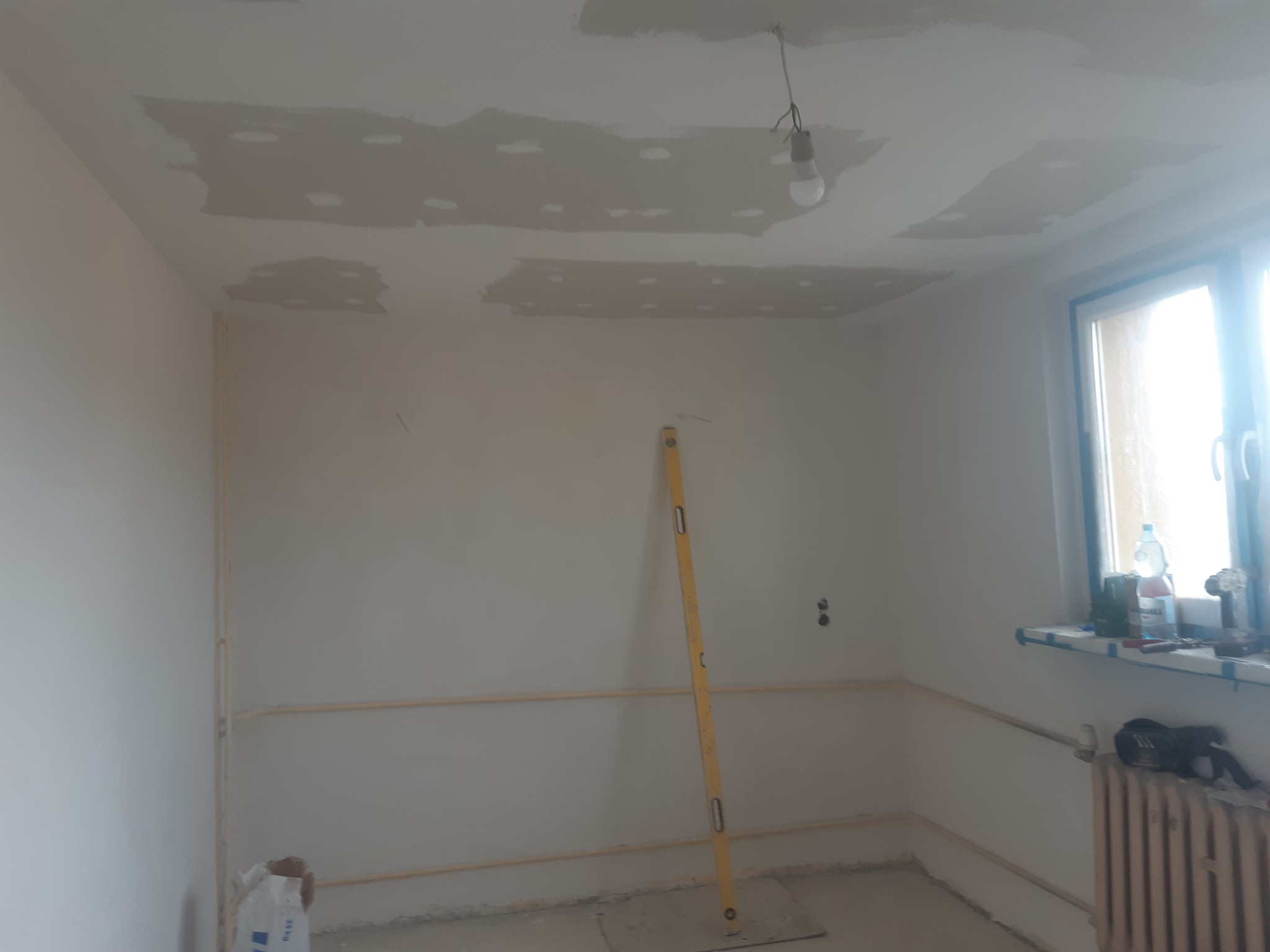 Pokój w trakcie prac wykończeniowych: szpachlowane fragmenty sufitu, poziomica oparta o ścianę, widoczne instalacje. Przy oknie narzędzia i grzejnik.