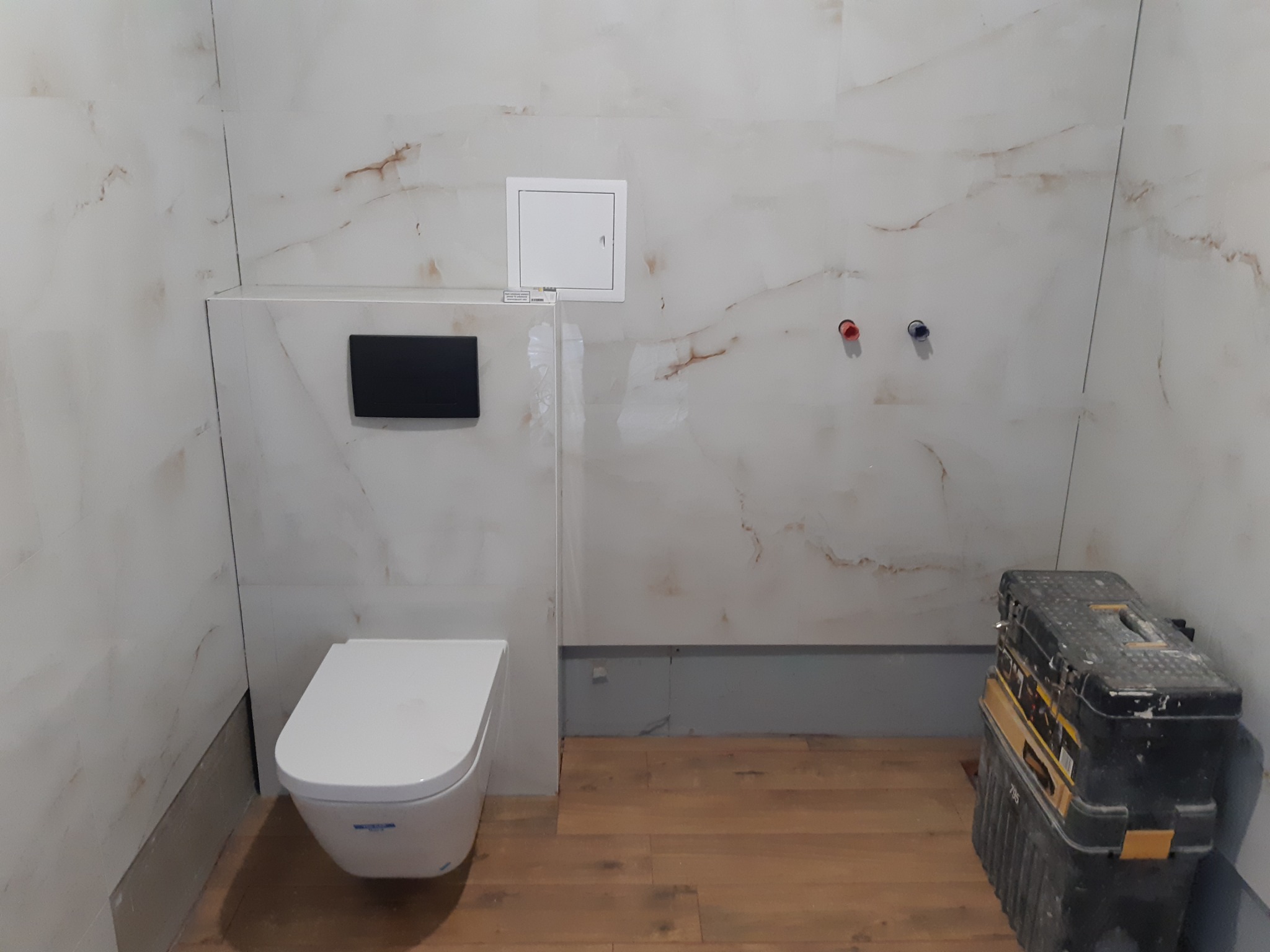 Nowoczesna toaleta wisząca w łazience z marmurowymi płytkami. Widoczna skrzynka narzędziowa, co sugeruje prace wykończeniowe. Podłoga wyłożona panelami.