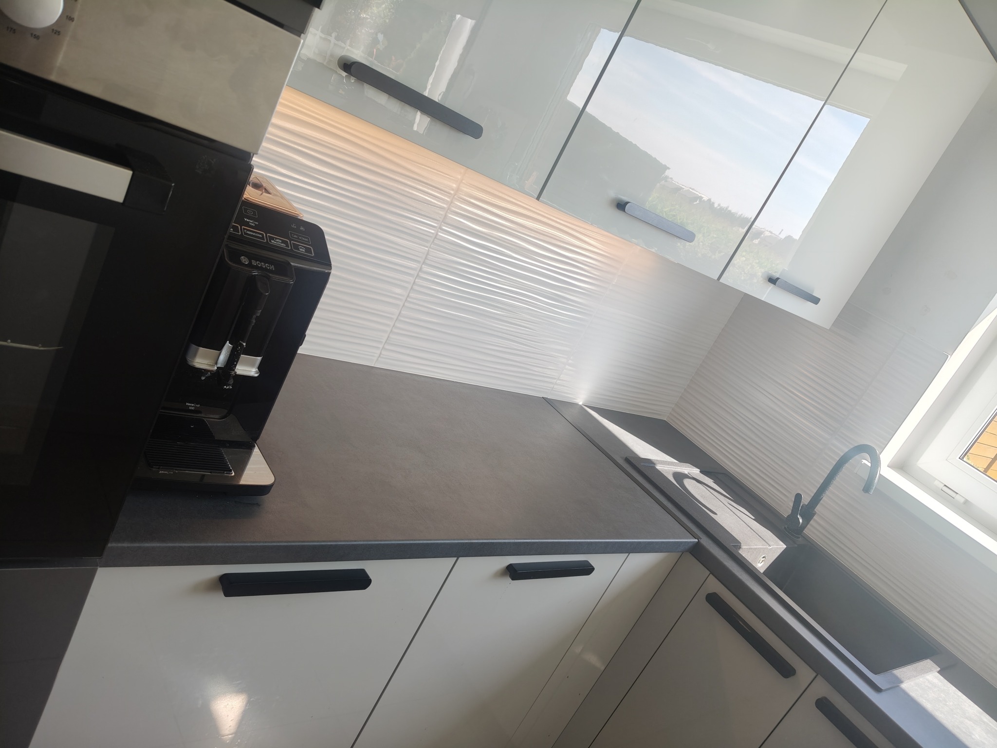 Nowoczesna, jasna kuchnia z białymi szafkami, szarym blatem i designerską armaturą. W tle okno wpuszczające naturalne światło, a na blacie ekspres do kawy marki Bosch.