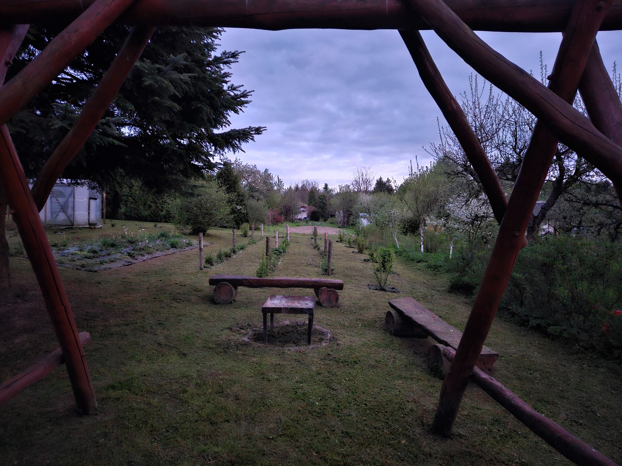 Widok na ogród z drewnianej konstrukcji. W tle drzewa, krzewy i trawnik. W centrum drewniane ławki i stolik. Widoczne ślady koszenia trawy.
