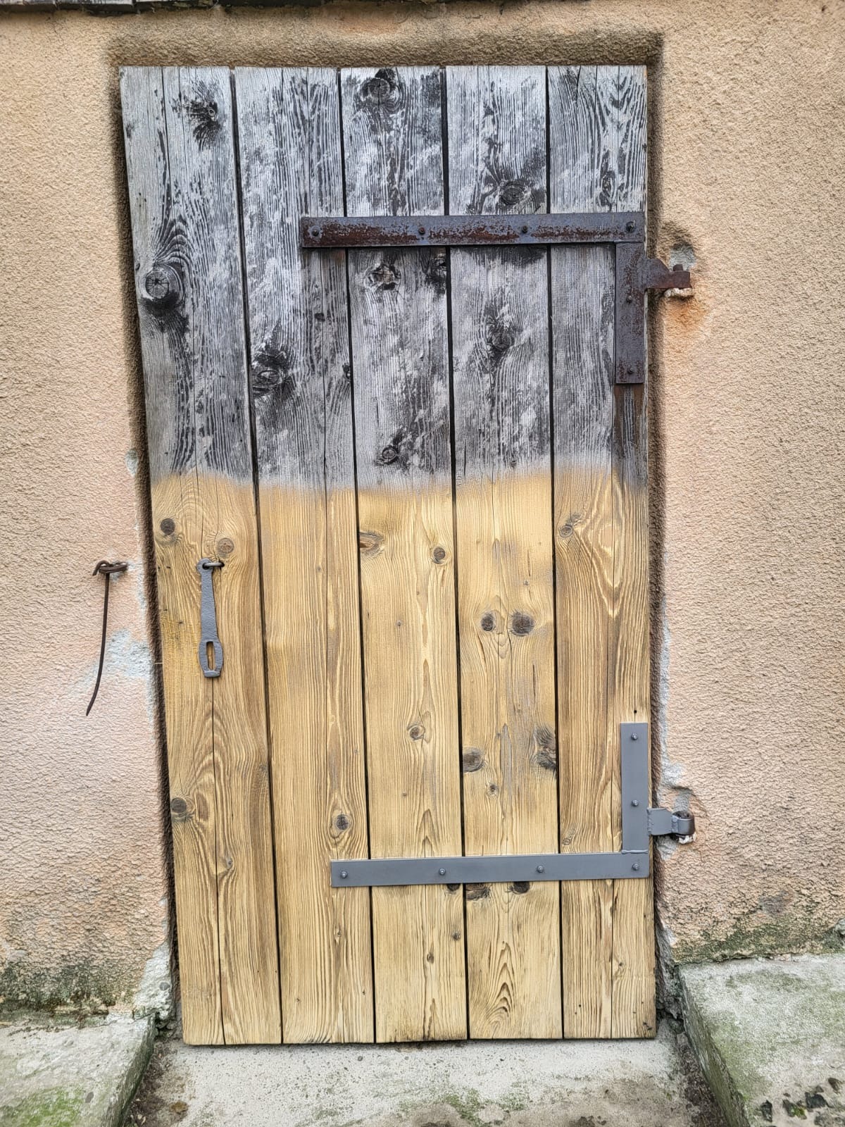 Drewniana brama z metalowymi okuciami, w połowie oczyszczona, ukazująca efekt odnowienia. Górna część szara, dolna w naturalnym odcieniu drewna.