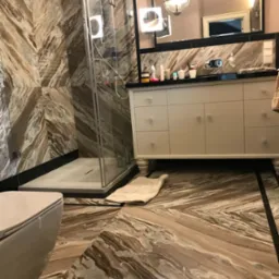 Elegancka łazienka z marmurowymi płytami na ścianach i podłodze, kabina prysznicowa ze szkła, biała szafka z umywalką i lustro z oświetleniem.