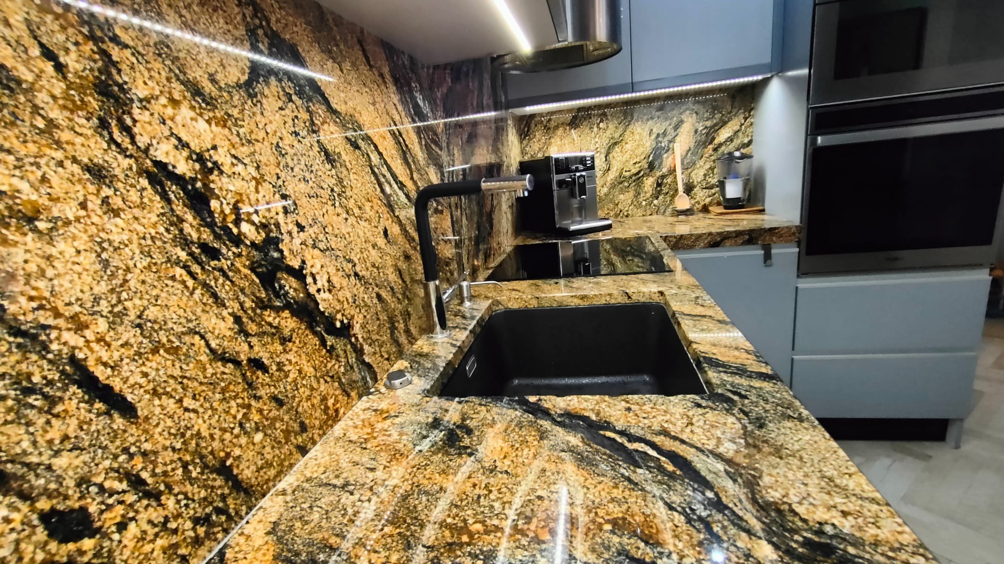 Kuchnia z blatem kamiennym o złoto-czarnym wzorze. Widoczny zlew, bateria, ekspres do kawy i oświetlenie LED pod szafkami. Ujęcie z bliska, podkreślające unikalny rysunek kamienia.