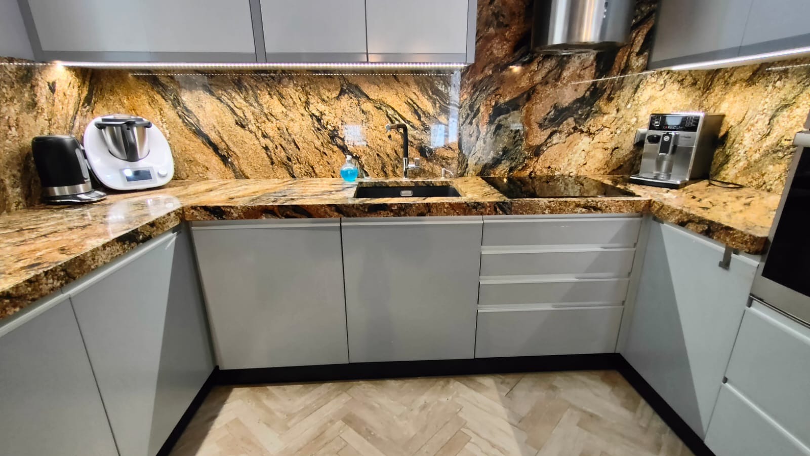 Kuchnia z blatem z kamienia o unikalnym, złotobrązowym wzorze. Uwagę przykuwa wysoki połysk blatu i dopasowany kolorystycznie panel nad blatem. Nowoczesne, szare szafki.