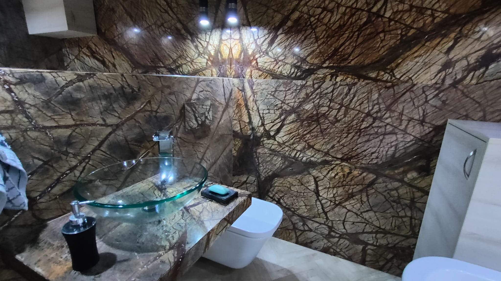 Łazienka z blatem kamiennym o wzorze drzewa, szklaną umywalką i ścianami z kamienia w tym samym wzorze. Nowoczesny design z oświetleniem punktowym.