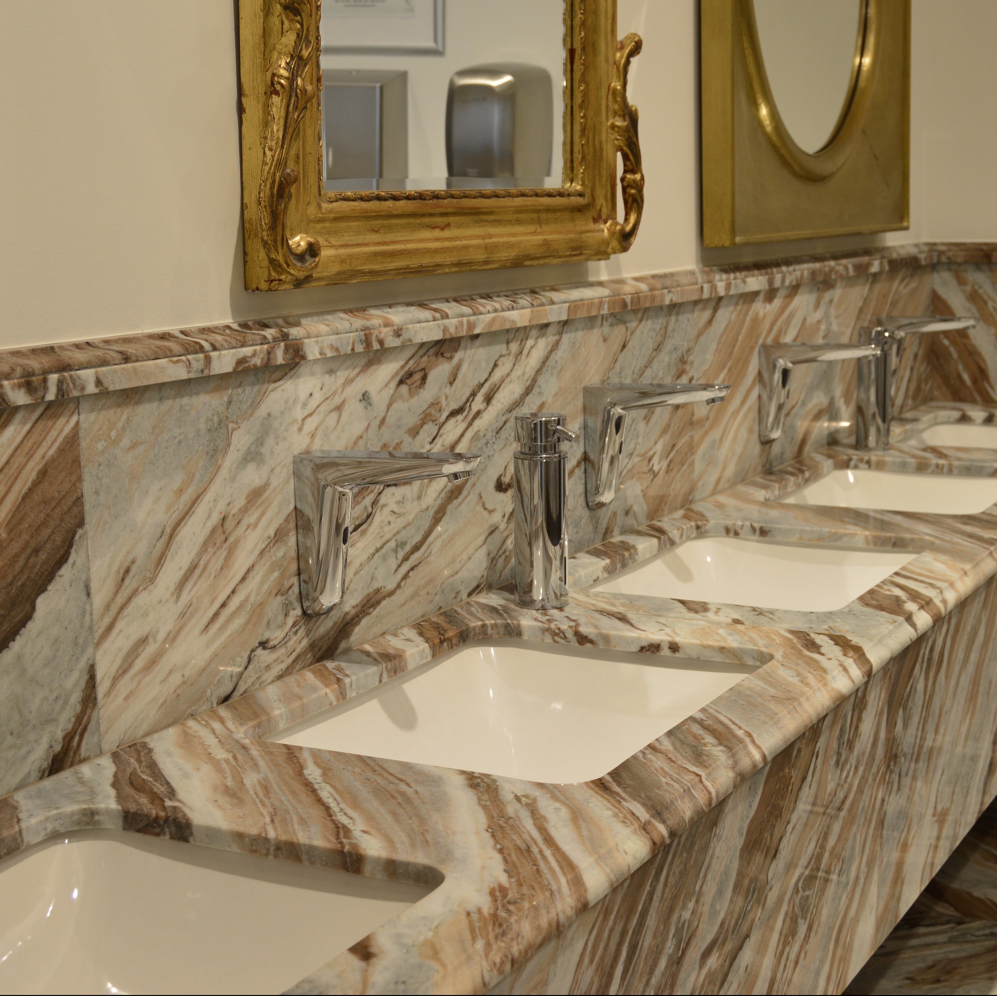Marmurowa umywalka z trzema białymi zlewami i chromowanymi bateriami, uzupełniona o złote ramy luster. Kamień o unikalnym wzorze.