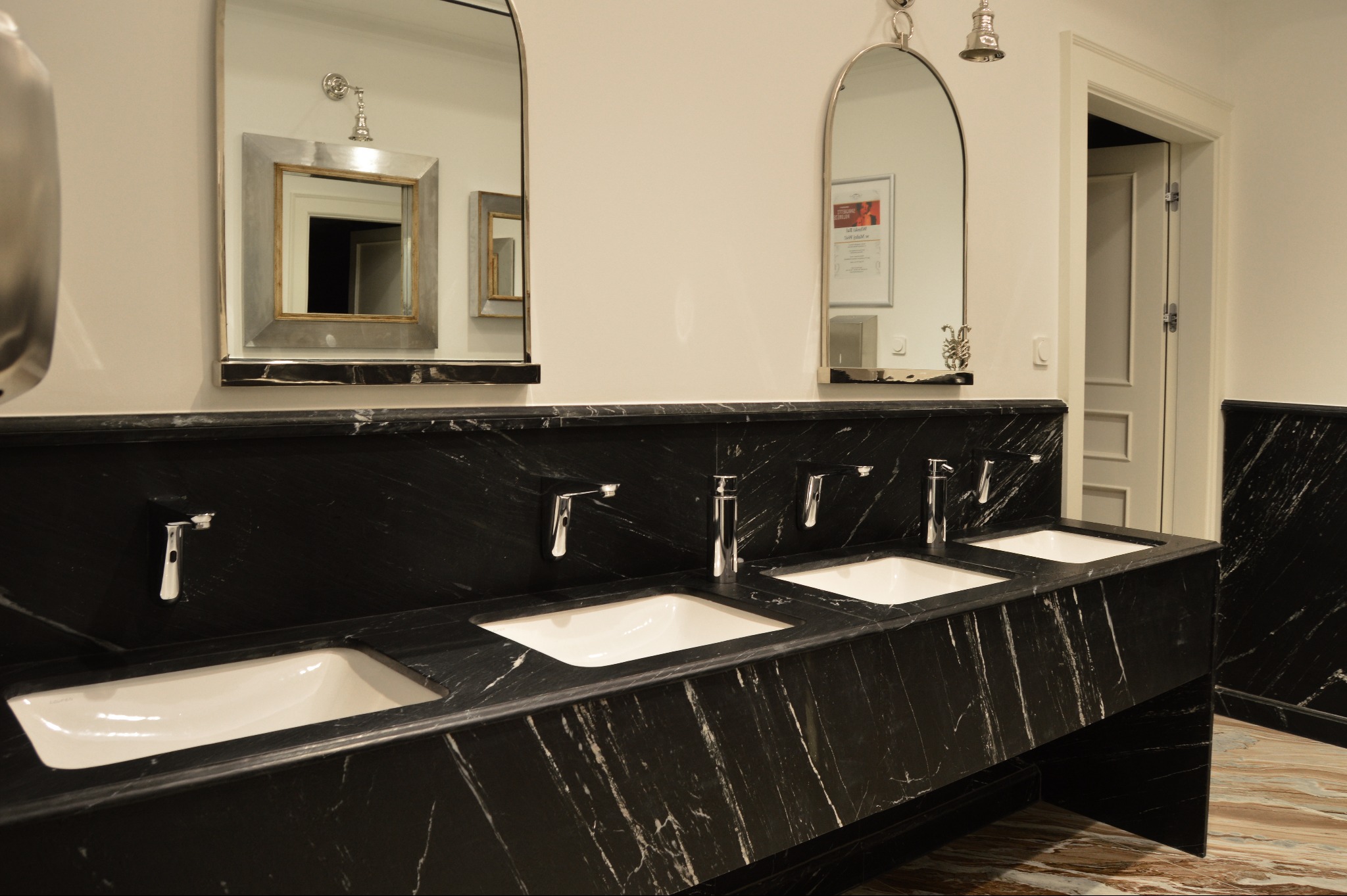 Czarny blat kamienny z trzema umywalkami i srebrnymi bateriami, zwieńczony lustrami o różnych kształtach. Widoczne marmurowe wykończenia ścian i podłoga.