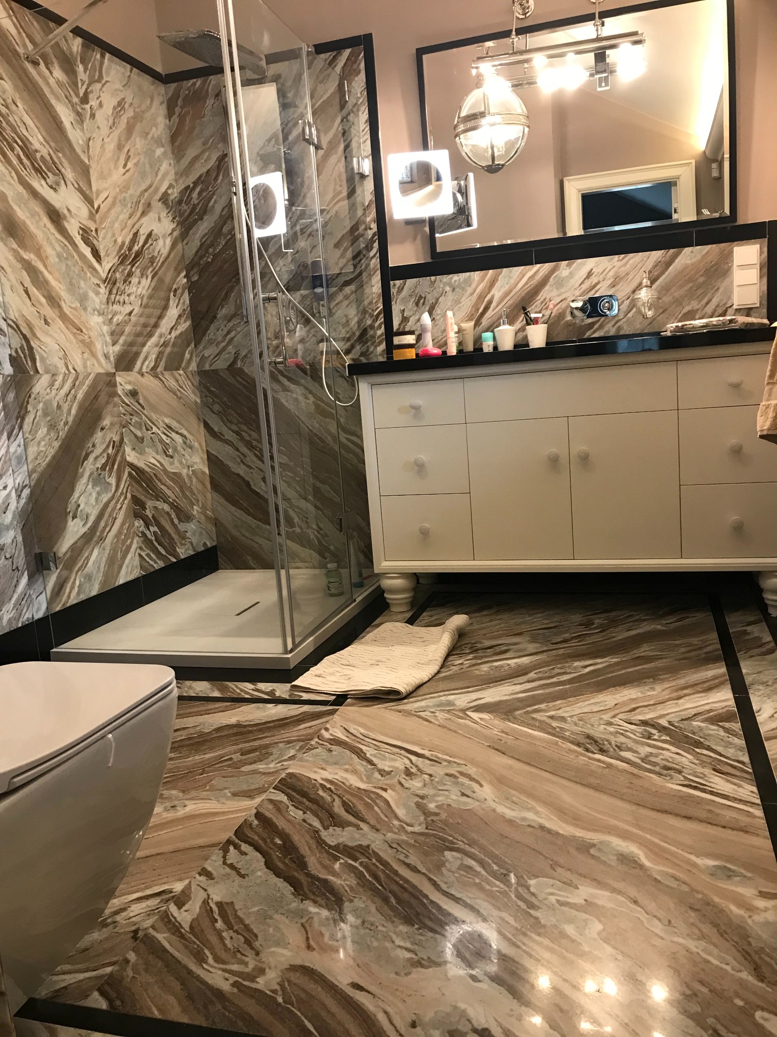 Elegancka łazienka z marmurowymi płytami na ścianach i podłodze, kabina prysznicowa ze szkła, biała szafka z umywalką i lustro z oświetleniem.