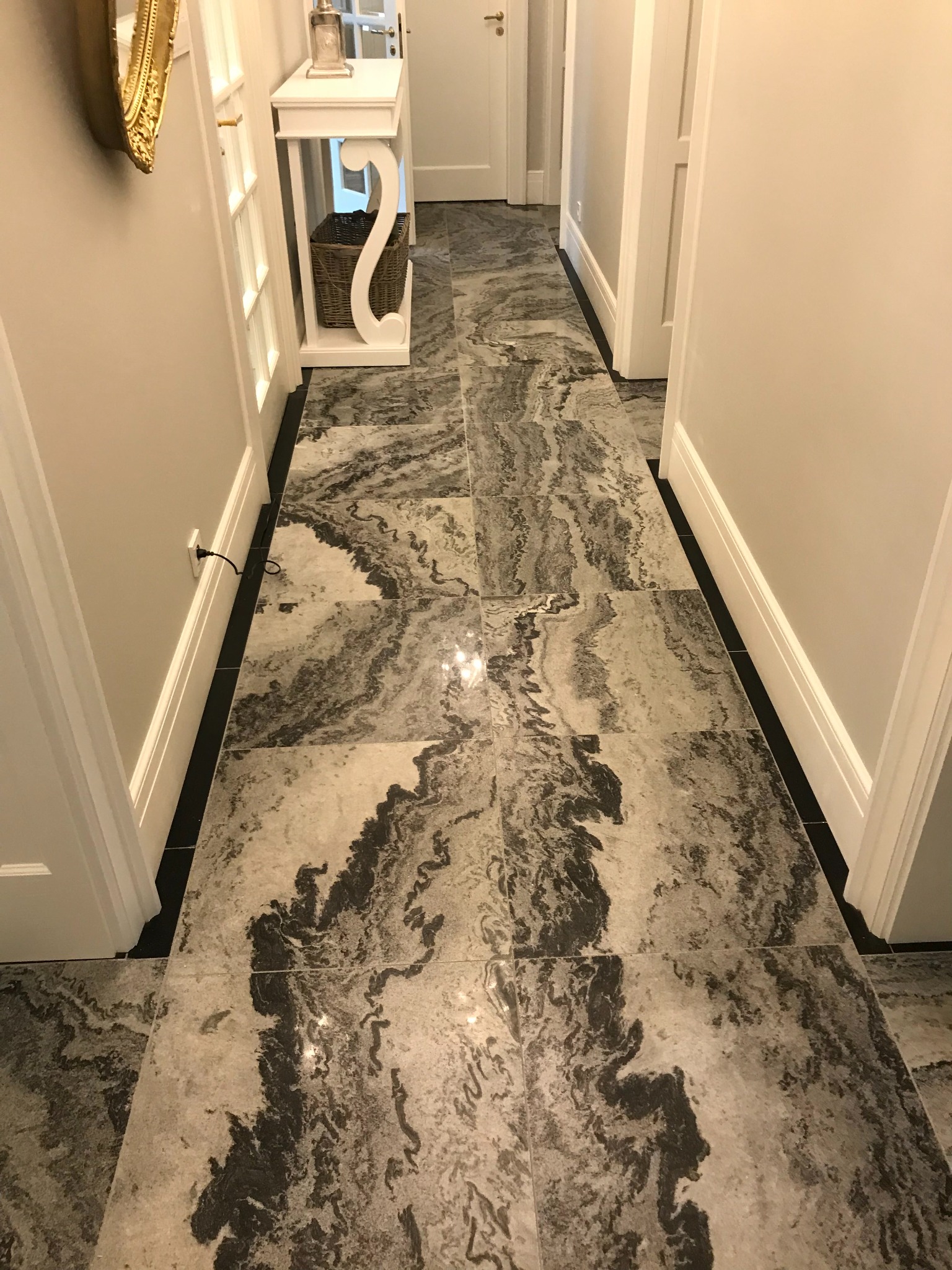 Korytarz z podłogą wyłożoną płytkami imitującymi kamień, o wzorze przypominającym marmur. Widoczne białe drzwi i listwy przypodłogowe, a także konsola z koszem.