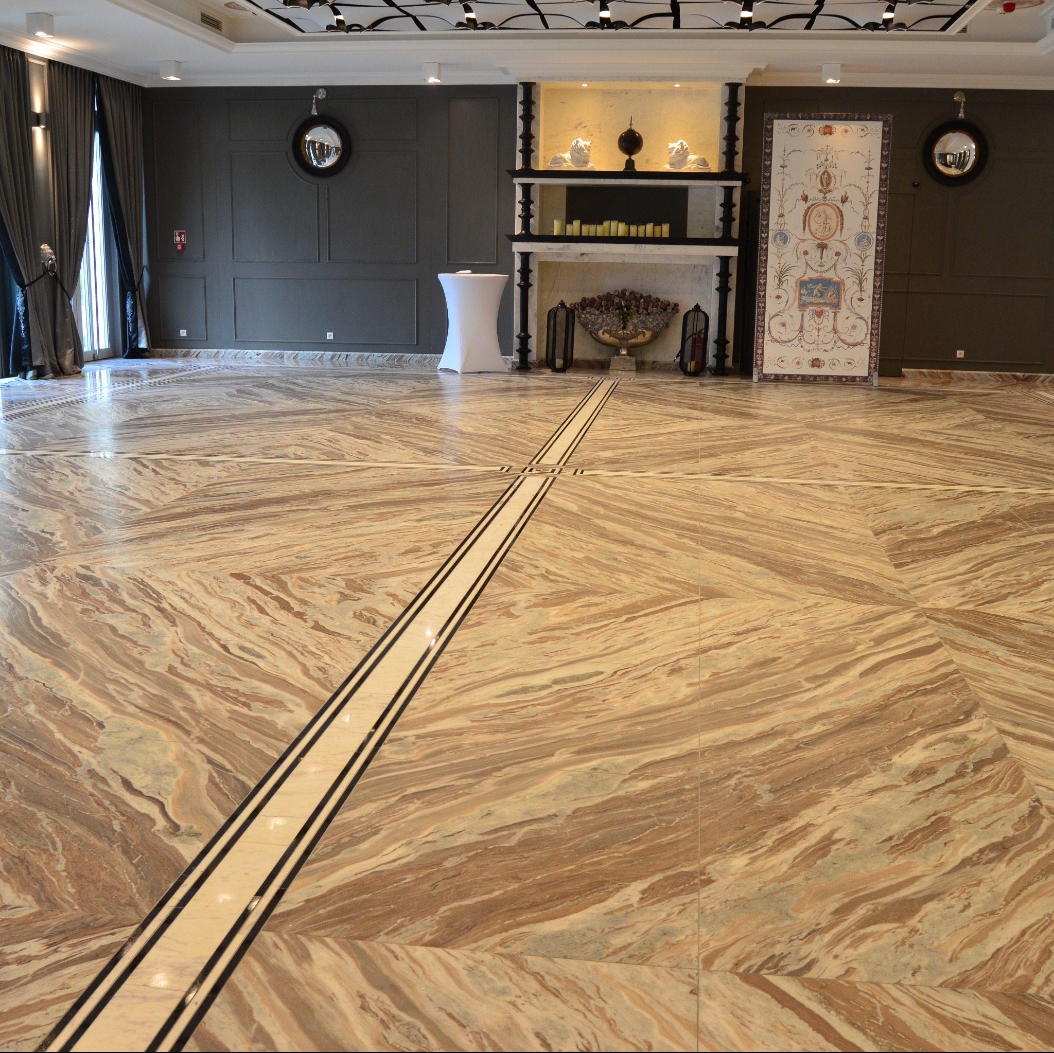 Elegancka posadzka z kamienia naturalnego o wzorze krzyża, zdobi wnętrze z szarą ścianą i dekoracyjnym kominkiem. Widoczne detale wykończenia i oświetlenie.