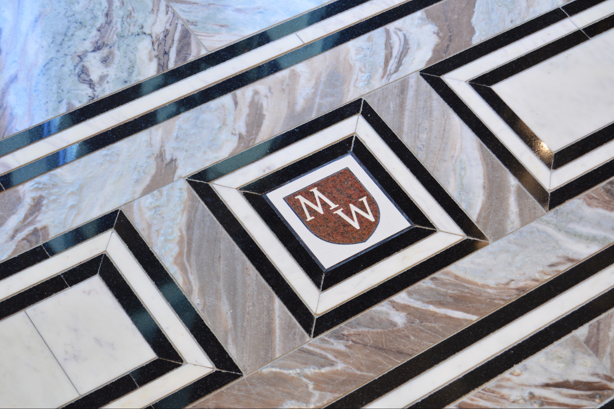 Wzorzysta kamienna posadzka z herbem 'MW' w centrum, ułożona z różnych rodzajów kamienia w odcieniach bieli, szarości, brązu i czerni. Geometryczny wzór z wyraźnymi liniami.