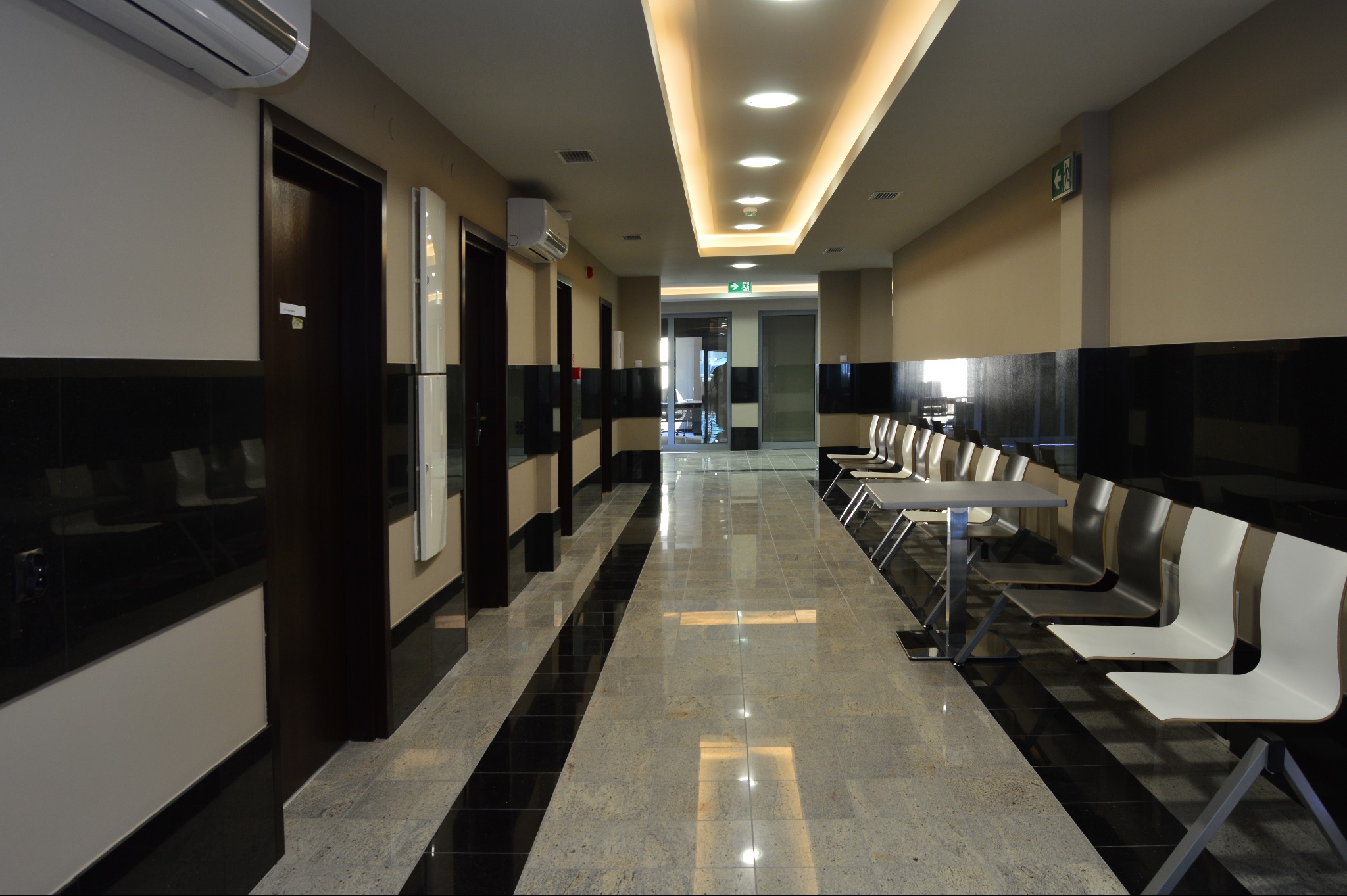Błyszcząca posadzka z kamienia w korytarzu z rzędem krzeseł i stolików. Widoczne czarne panele ścienne i oświetlenie sufitowe.