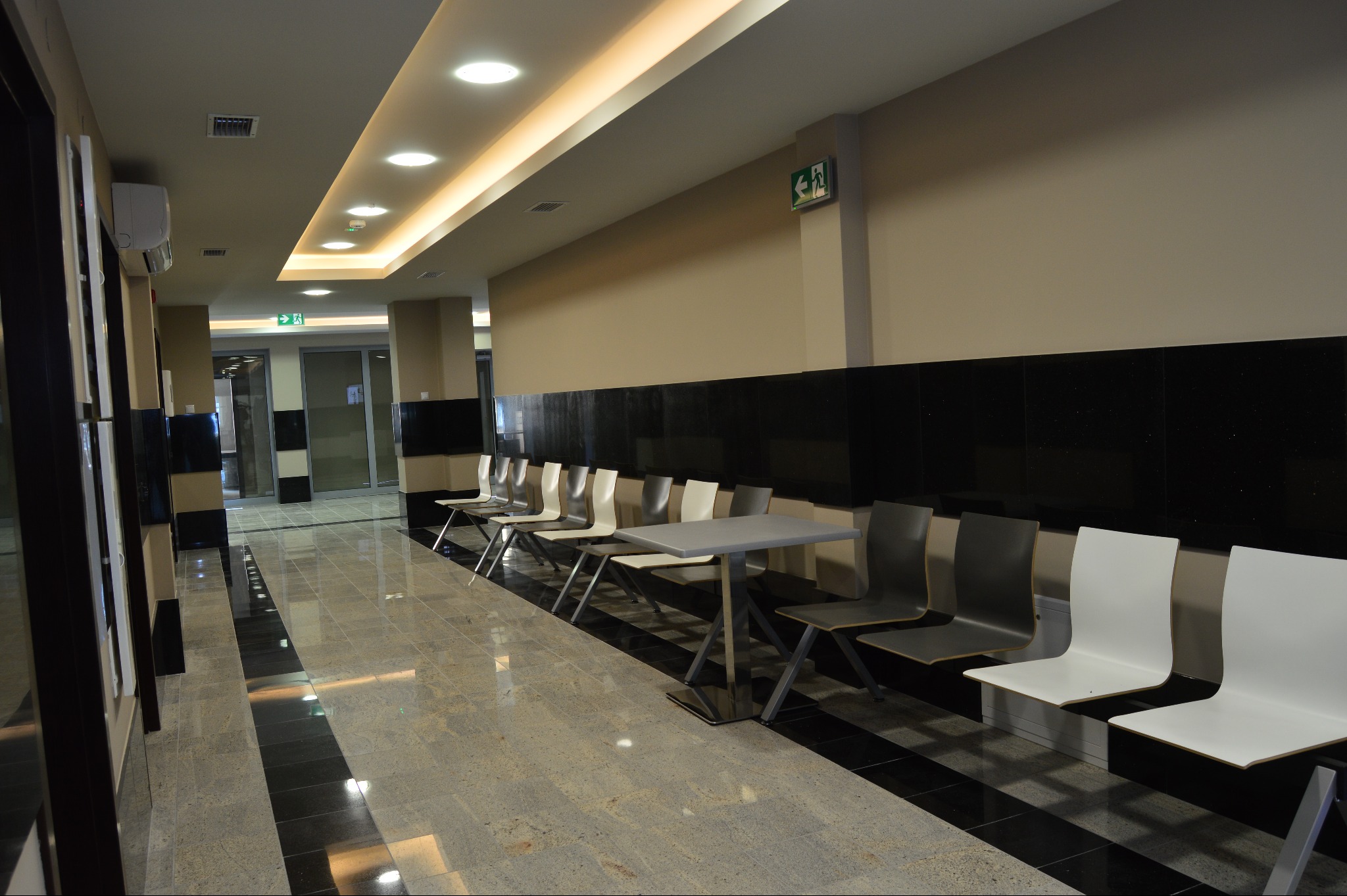 Hol we wnętrzu budynku z polerowaną posadzką i rzędem krzeseł. Ściany wykończone panelami z czarnego kamienia. Nowoczesne oświetlenie sufitowe.