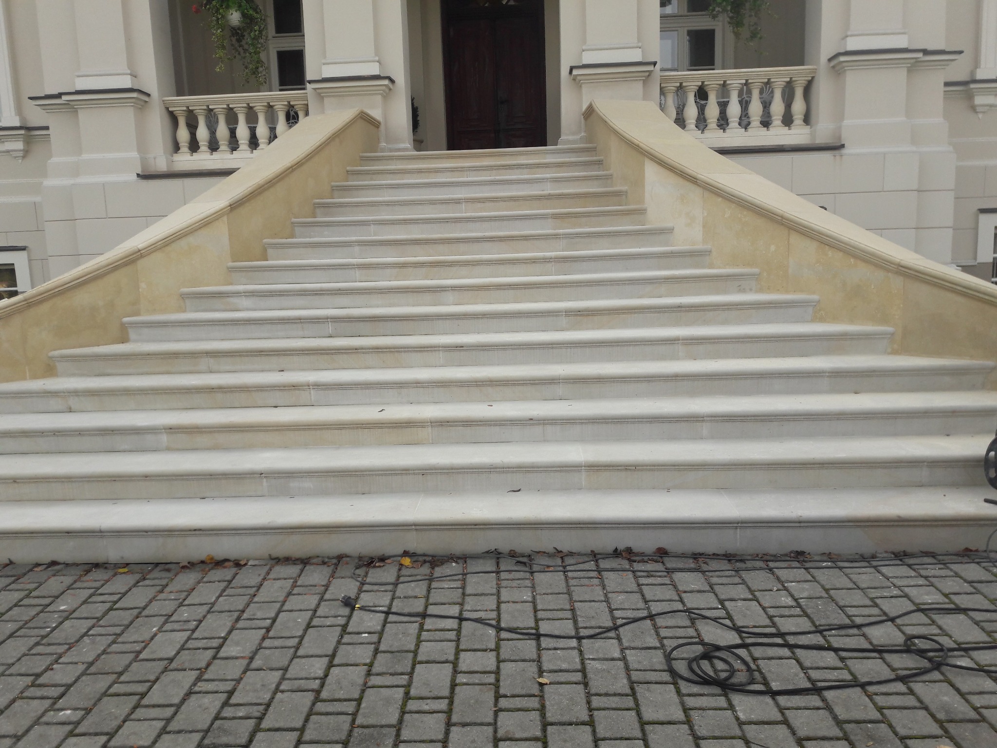 Jasne, kamienne schody zewnętrzne z balustradami, prowadzące do budynku z ciemnymi drzwiami. Na brukowanym podłożu leży czarny kabel.
