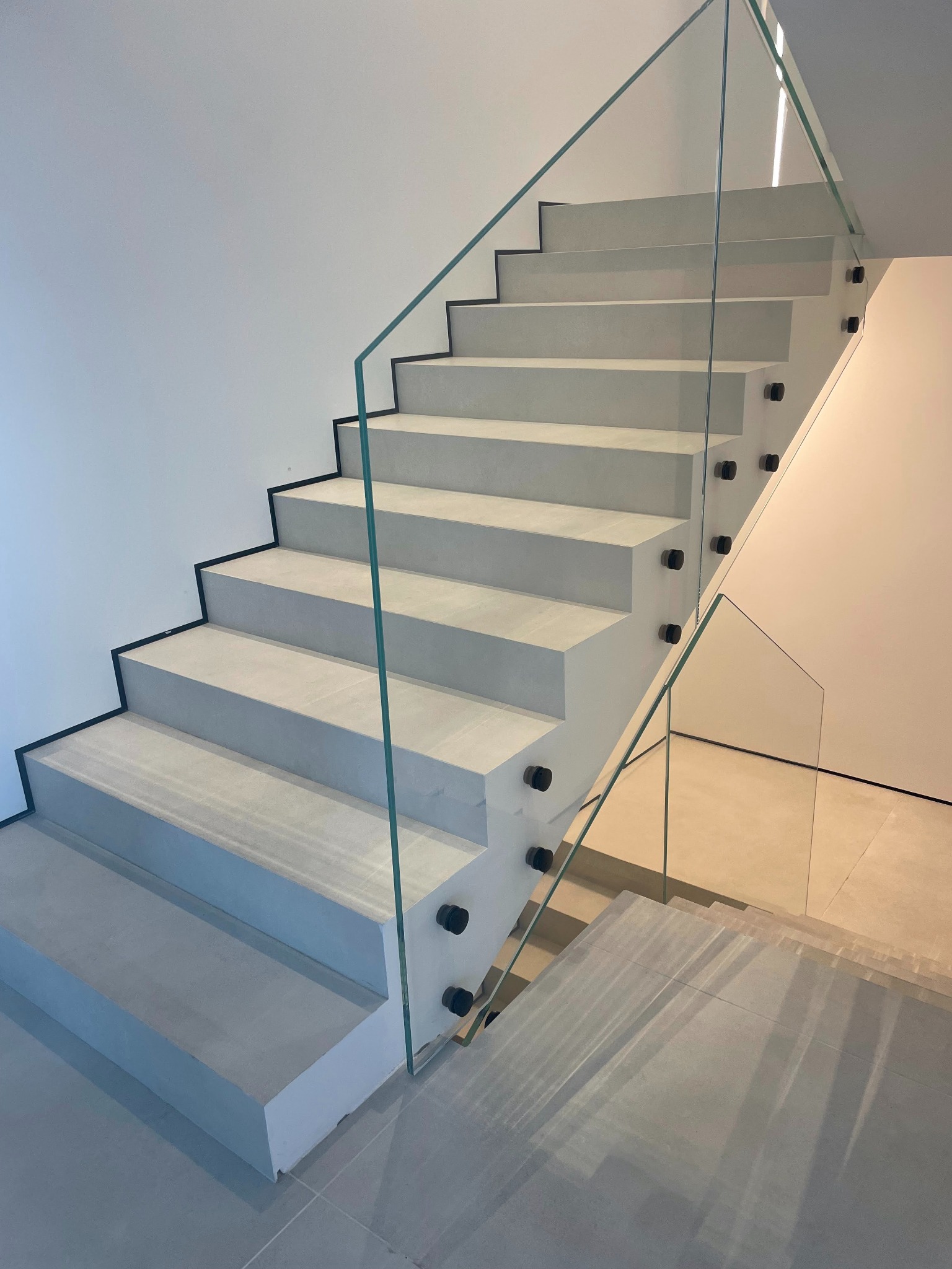 Nowoczesne schody z szarego kamienia z szklaną balustradą, mocowaną czarnymi uchwytami. Minimalistyczny design, jasne wnętrze.