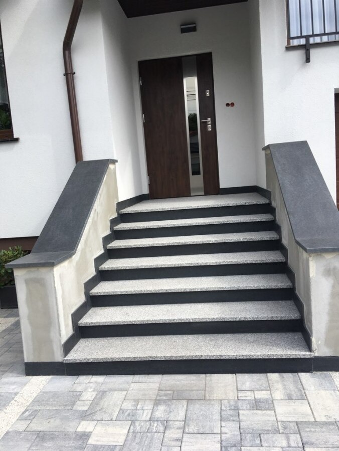 Zewnętrzne schody kamienne prowadzące do domu, z ciemnoszarymi obrzeżami i jasnoszarym wypełnieniem stopni. Widoczne wykończenie elewacji i fragment okna.