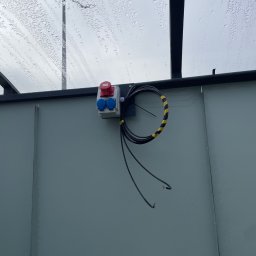 KON-TECH ADRIAN KONIECZNY - Zewnętrzna skrzynka elektryczna z gniazdami (czerwone i niebieskie) zamontowana na szarej ścianie pod zadaszeniem z kroplami deszczu. Kabel zwinięty i przymocowany taśmą.