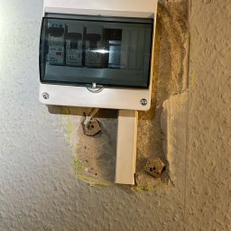 KON-TECH ADRIAN KONIECZNY - Zamontowana skrzynka elektryczna z widocznymi bezpiecznikami na nierównej, szarej ścianie. Widoczne ślady po wcześniejszych instalacjach i niedokładne wykończenie.
