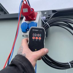 KON-TECH ADRIAN KONIECZNY - Panel elektryczny z gniazdami siłowymi, sterowany pilotem Voltcraft VC-32. Widoczne kable i instalacja. Ujęcie z bliska, z ręką trzymającą pilot.