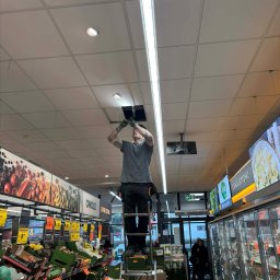 KON-TECH ADRIAN KONIECZNY - Elektryk na drabinie w supermarkecie, montuje oświetlenie sufitowe. Widoczne regały z owocami i chłodziarki z gotowymi daniami. Praca w przestrzeni handlowej.