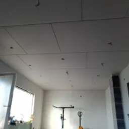 Wnętrze w trakcie montażu płyt kartonowo-gipsowych na suficie, widoczne narzędzia i elementy instalacji elektrycznej. Okno z lewej strony, surowy stan wykończenia.