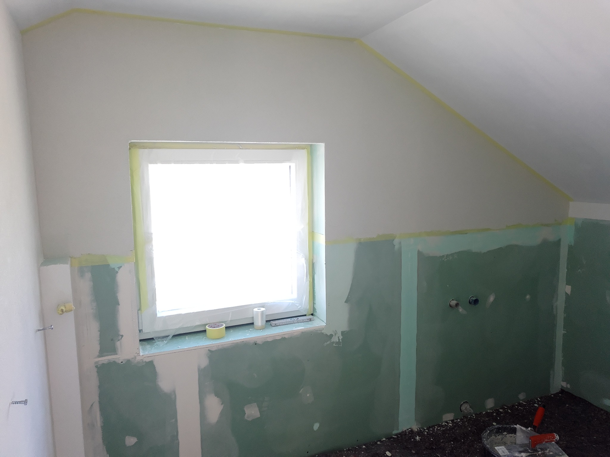 Poddasze w trakcie wykańczania: ściany z płyt gipsowo-kartonowych, częściowo zaszpachlowane, okno zabezpieczone taśmą malarską, widoczne narzędzia i akcesoria.