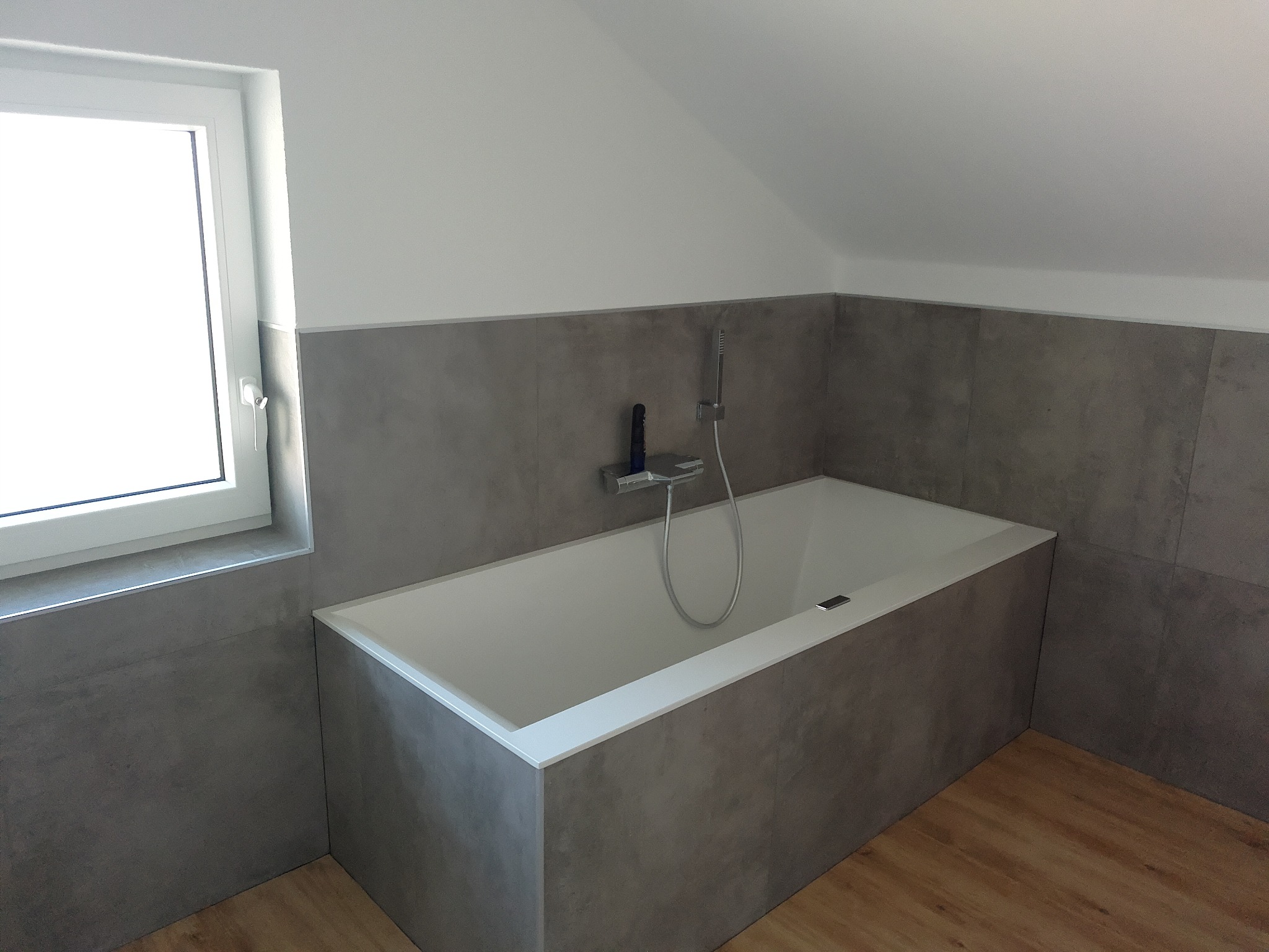 Nowoczesna wanna obudowana szarą płytką w łazience na poddaszu, z oknem po lewej stronie. Minimalistyczny design, drewniana podłoga.