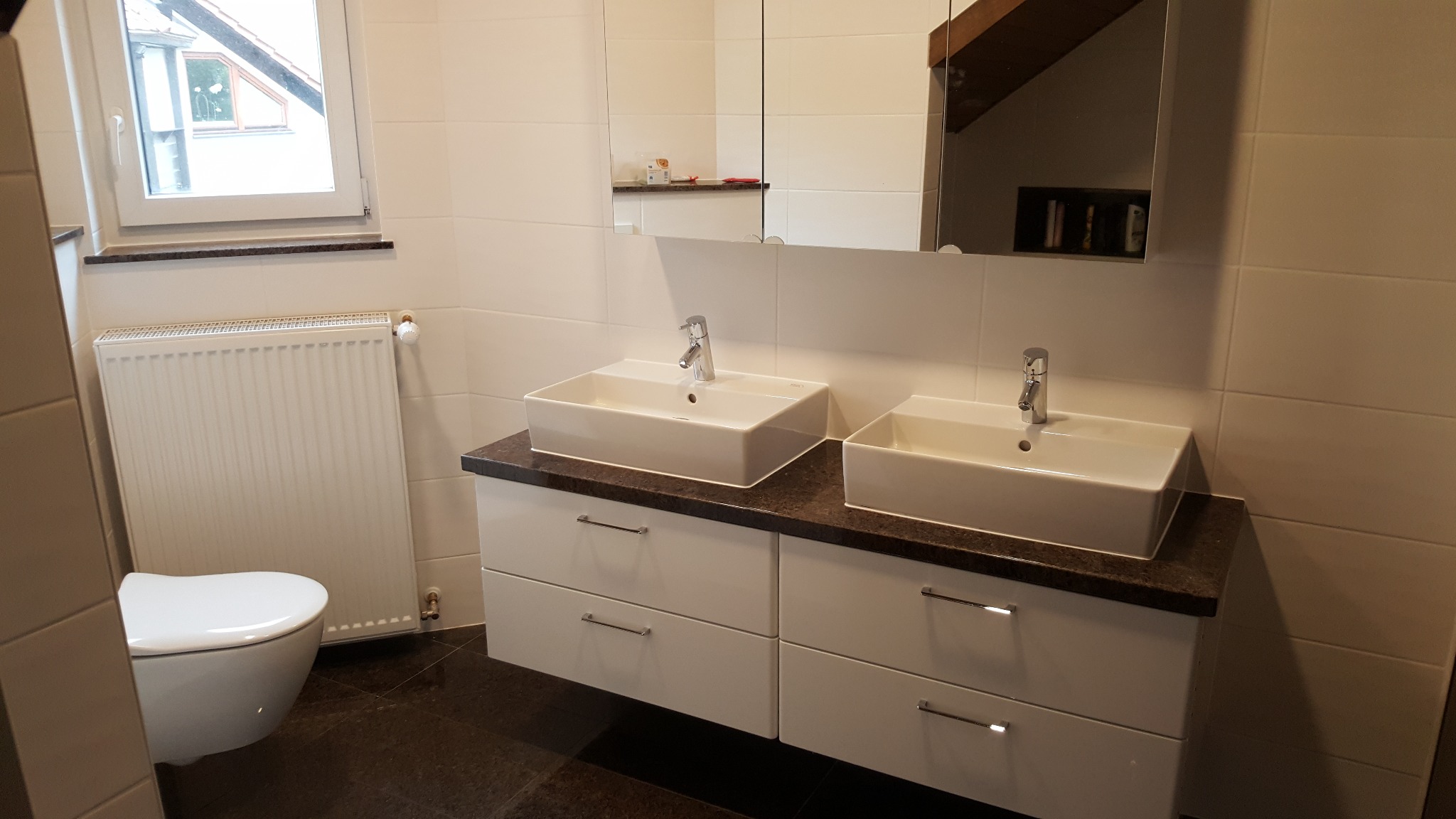 Nowoczesna łazienka z dwoma prostokątnymi umywalkami nablatowymi, białymi szafkami z szufladami i dużym lustrem. Widoczny fragment toalety i grzejnik.