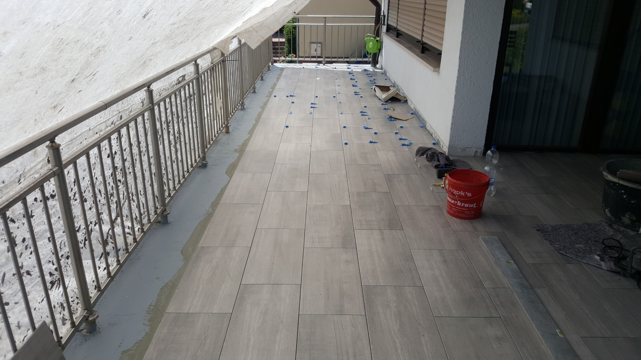 Układanie płytek na balkonie z metalową barierką. Widoczne narzędzia, wiadro i krzyżyki dystansowe. Jasne, naturalne światło. Częściowo zadaszony.