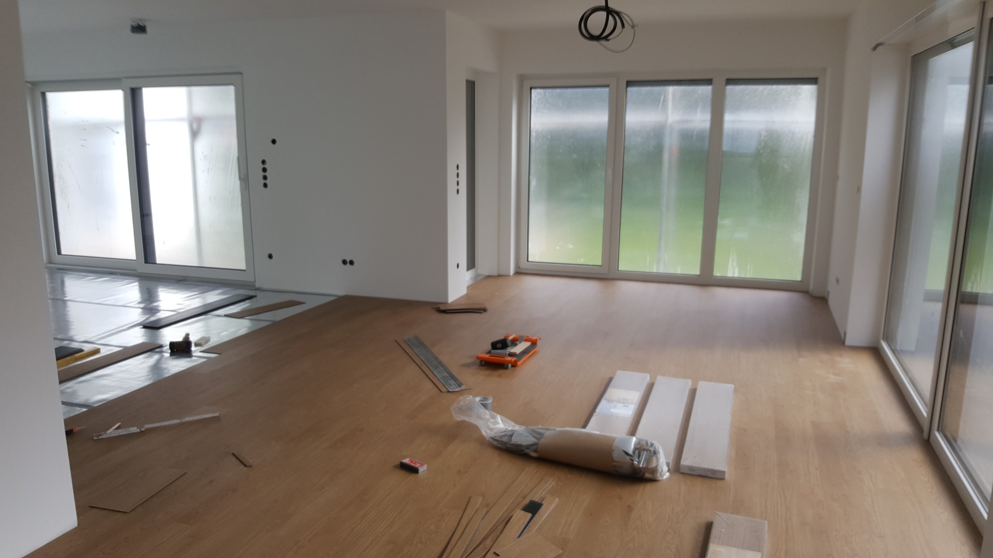 Układanie paneli podłogowych w jasnym wnętrzu z dużymi oknami. Widoczne narzędzia i materiały budowlane, częściowo ułożona podłoga.