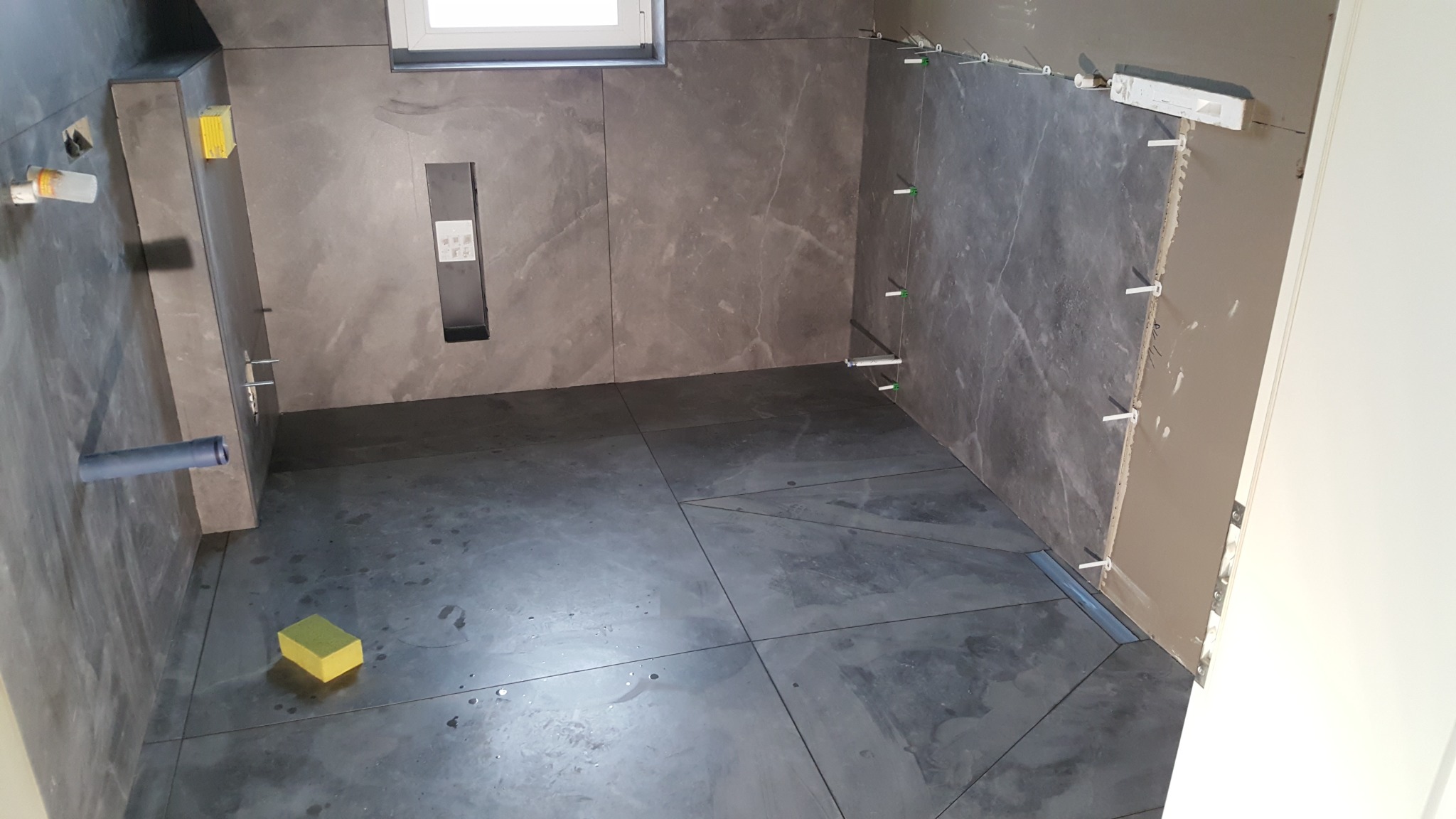 Wnętrze łazienki w trakcie wykańczania: ułożone ciemne płytki podłogowe i częściowo na ścianach, widoczne elementy instalacji, poziomowanie płytek, gąbka.
