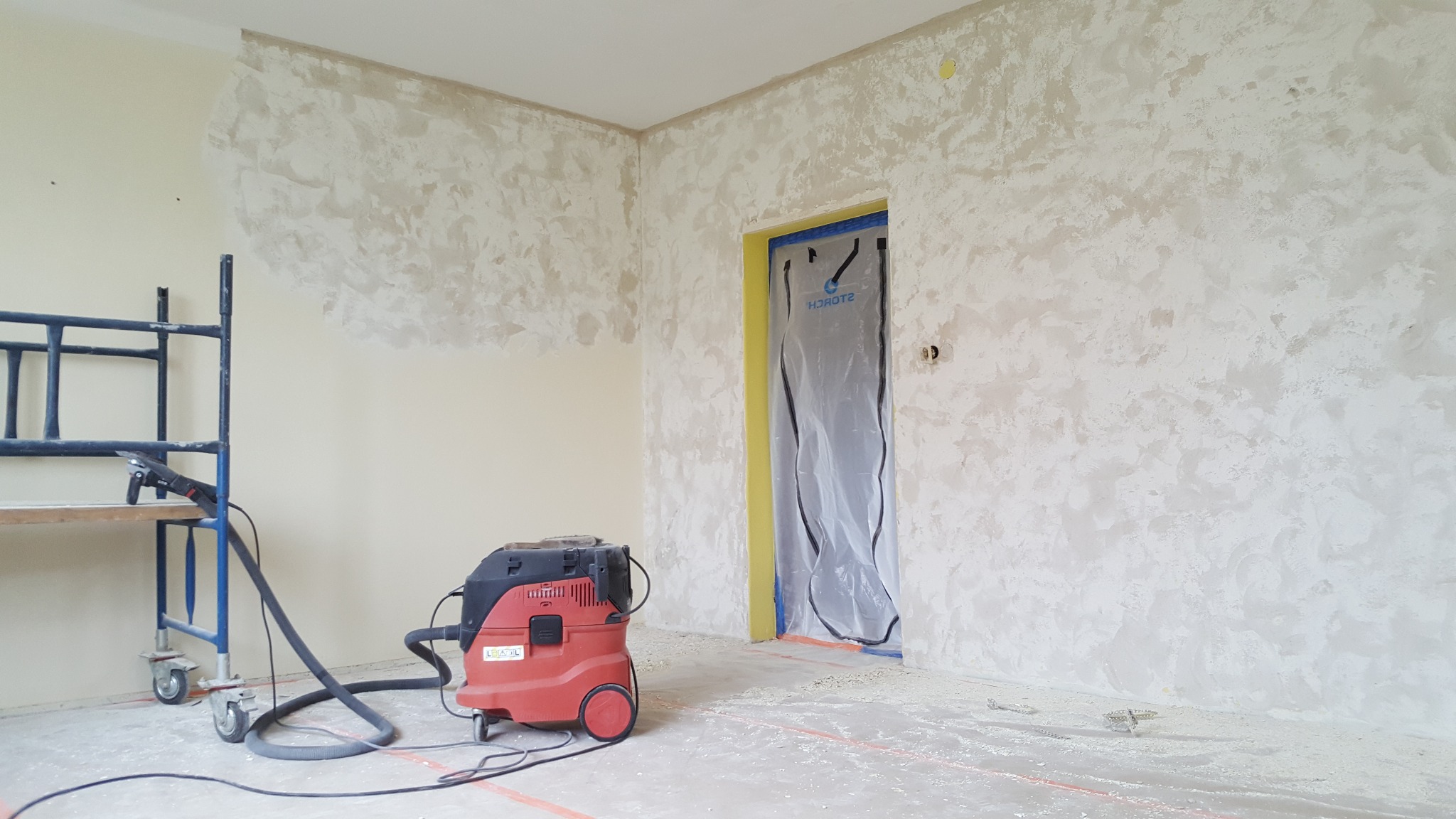 Pokój w trakcie tynkowania dekoracyjnego. Widoczne rusztowanie, odkurzacz przemysłowy i zabezpieczone folią drzwi. Ściany pokryte częściowo tynkiem.