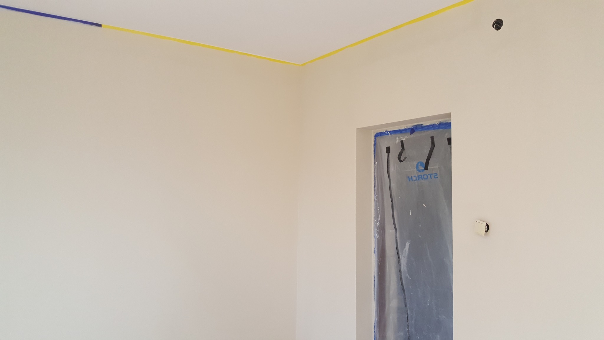Przygotowane do malowania wnętrze: świeżo pomalowane ściany w jasnym odcieniu, zabezpieczone taśmą malarską krawędzie sufitu i folią otwór drzwiowy.