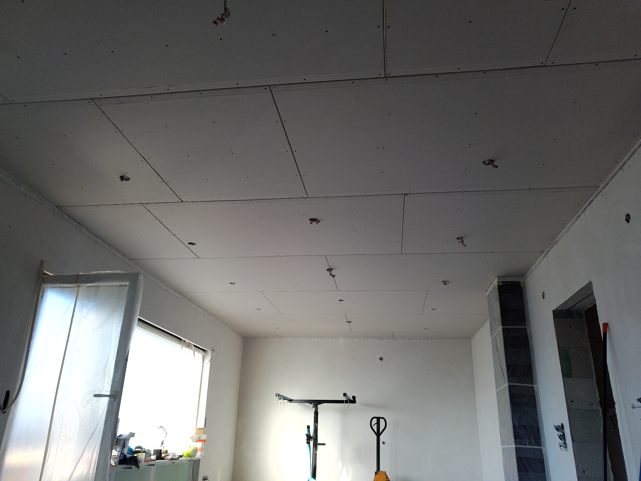 Wnętrze w trakcie montażu płyt kartonowo-gipsowych na suficie, widoczne narzędzia i elementy instalacji elektrycznej. Okno z lewej strony, surowy stan wykończenia.