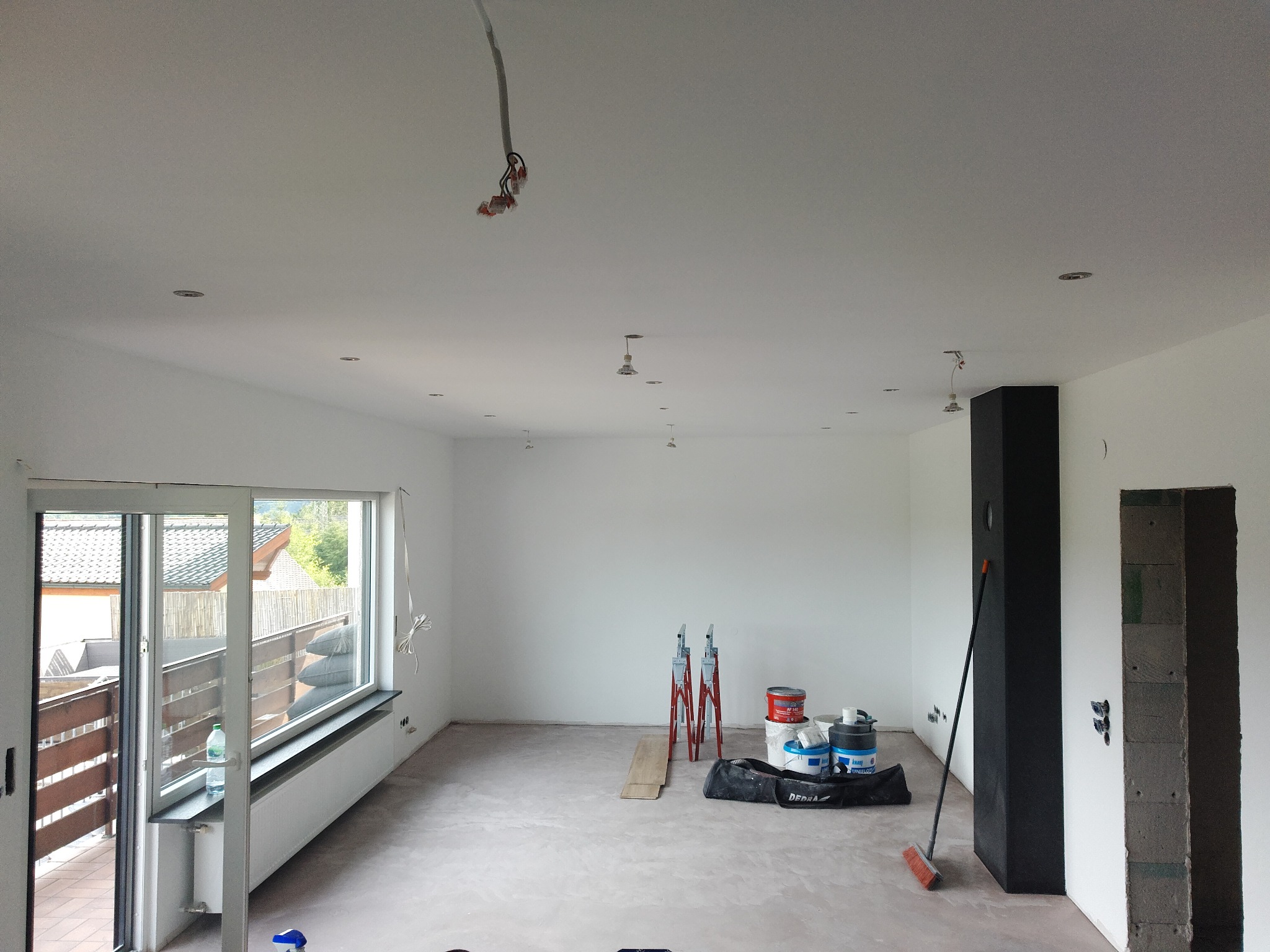 Wnętrze w trakcie remontu: białe ściany, surowa podłoga, drabiny malarskie, puszki z farbą i odsłonięte przewody elektryczne pod sufitem. Widok na balkon.