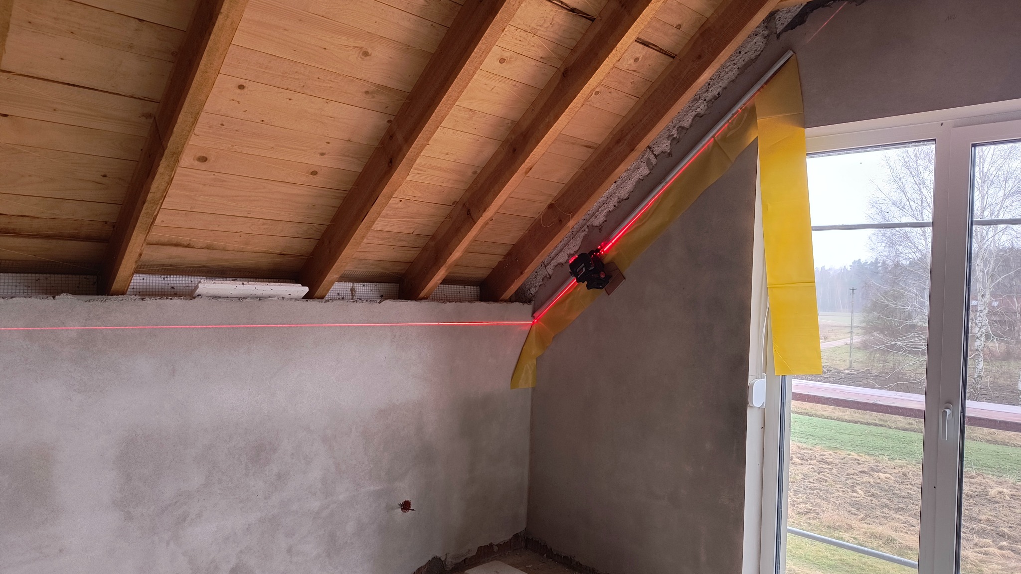 Skośne poddasze w trakcie tynkowania, widoczny laser krzyżowy wyznaczający poziom, drewniana konstrukcja dachu i okno z widokiem na zewnątrz.
