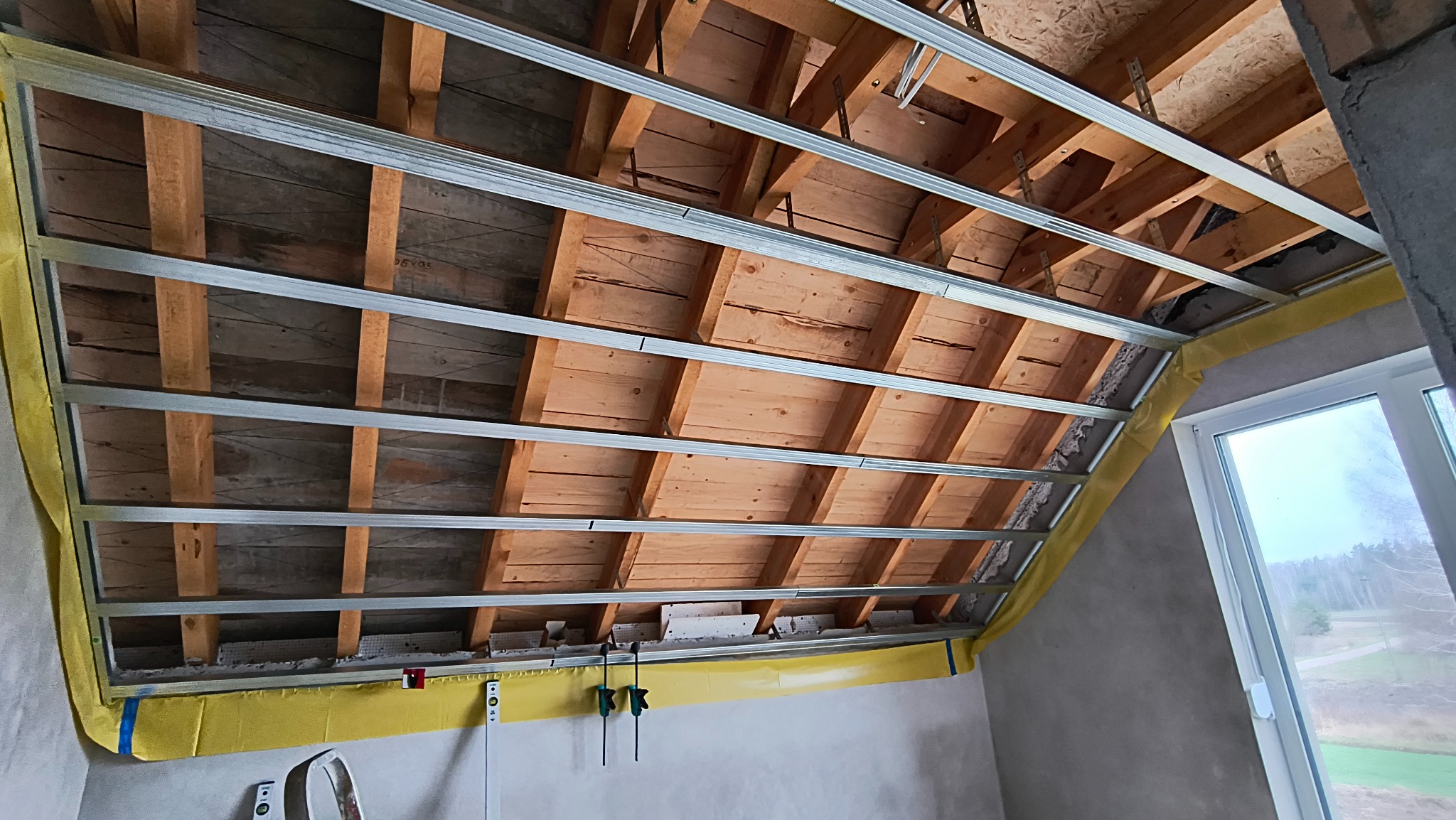 Skośny sufit z drewnianą konstrukcją i metalowym stelażem pod płyty gipsowe, zabezpieczony żółtą folią. Widok na okno i krajobraz za nim.