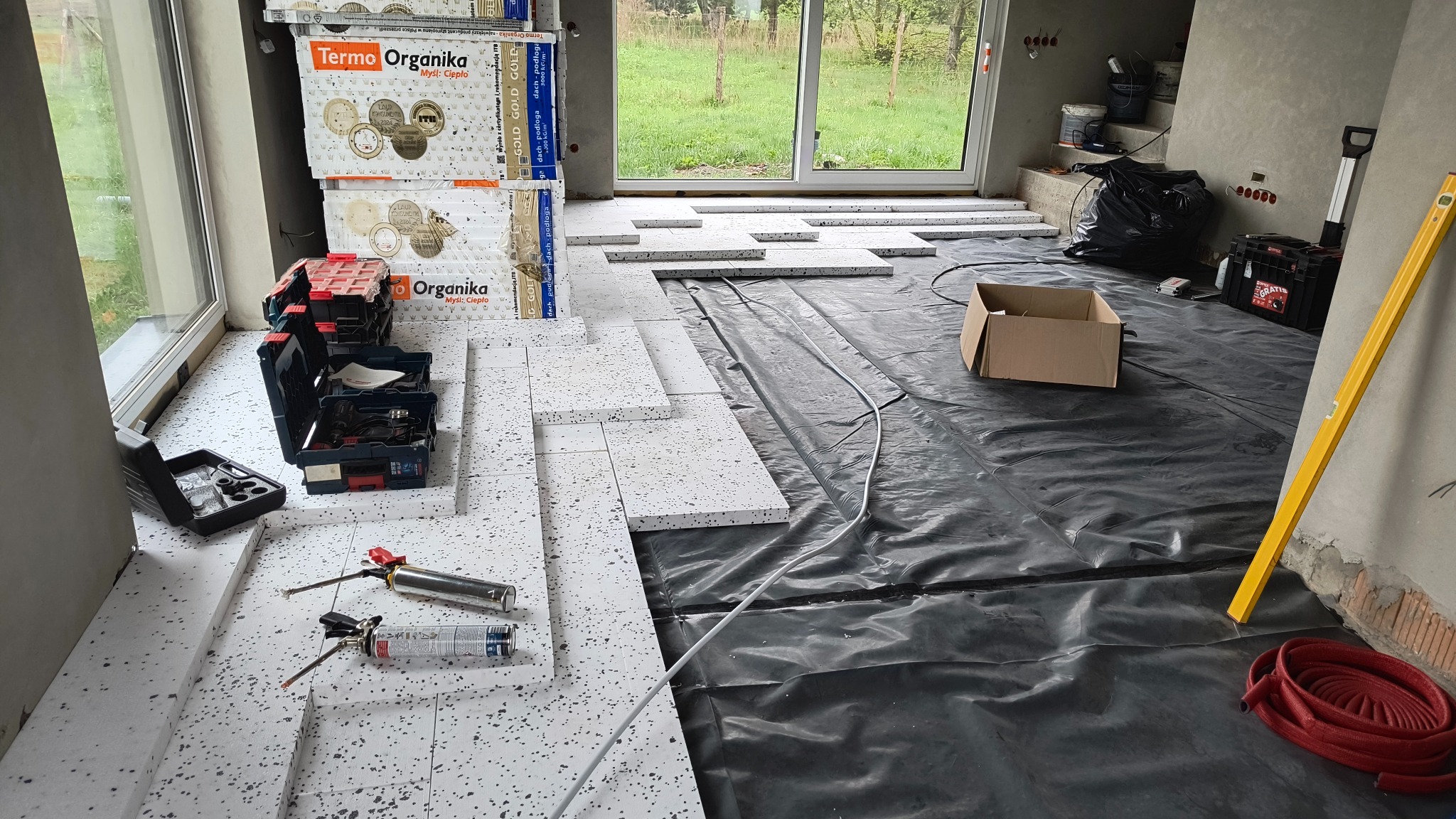Montaż izolacji podłogowej z płyt styropianowych na folii, widoczne narzędzia i materiały budowlane. Etap prac wykończeniowych w nowym budynku.