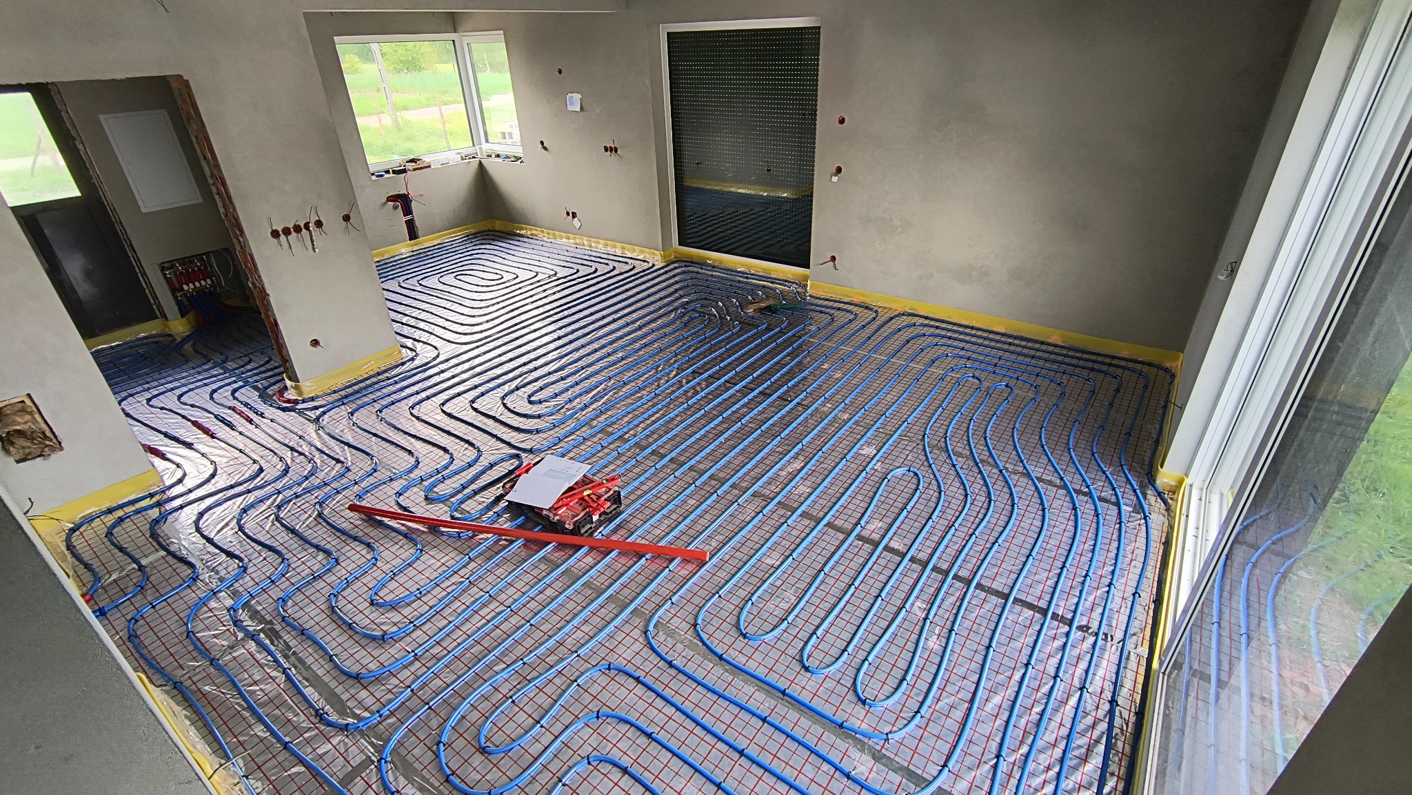 Montaż ogrzewania podłogowego w Brzezinkach. Niebieskie rurki ułożone na folii, widoczne narzędzia i surowe ściany. Etap instalacji przed wylaniem posadzki.