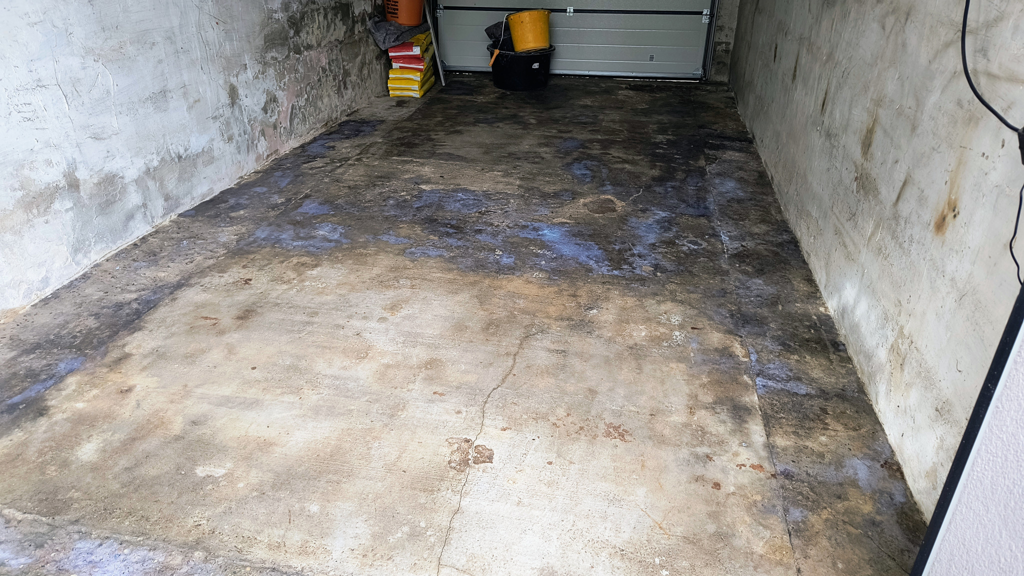 Surowy beton w garażu w Brzezinkach przed remontem. Widoczne ślady wilgoci i zabrudzenia na podłodze i ścianach. W tle wiaderka i worki z materiałami budowlanymi.