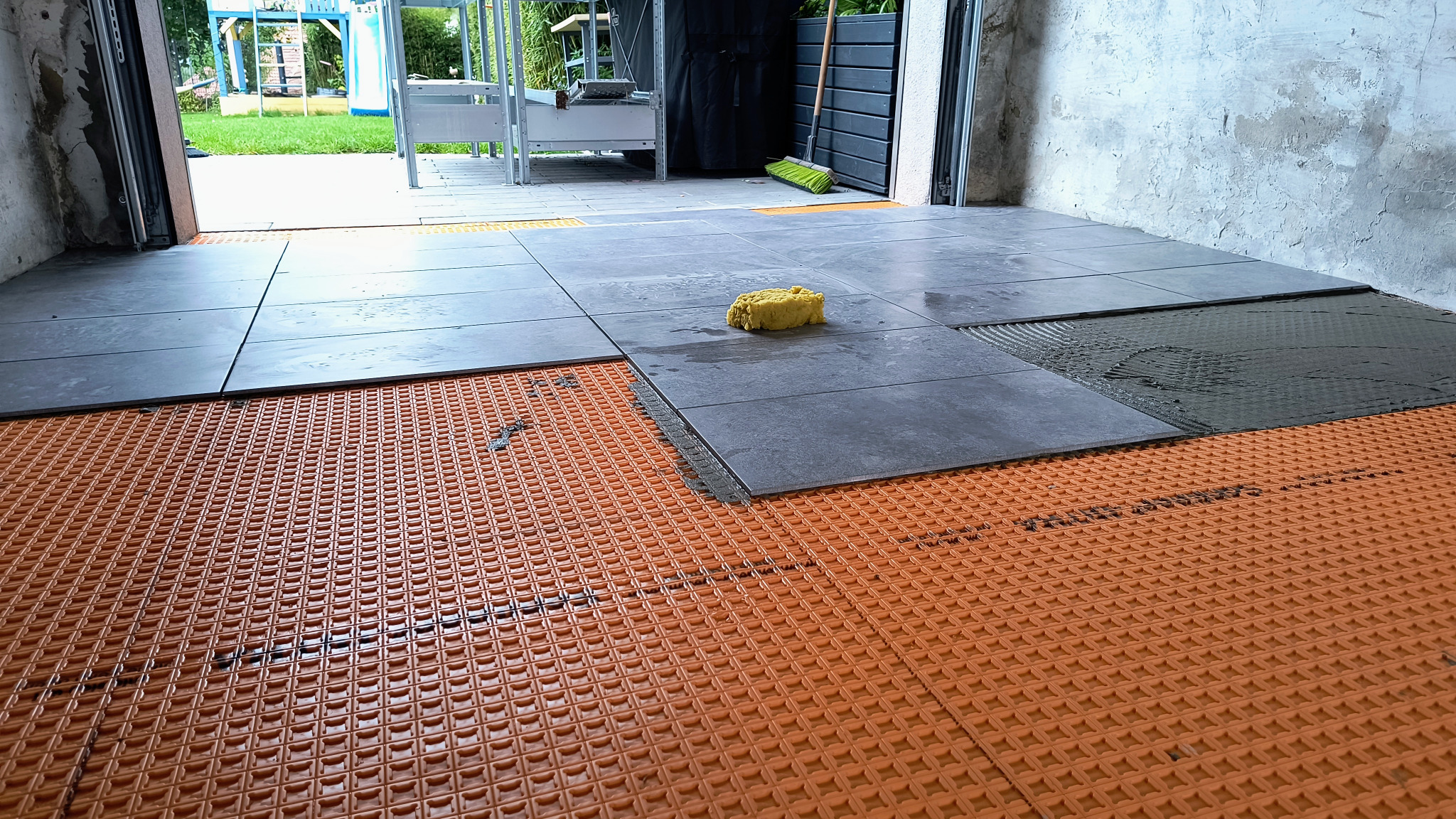 Układanie szarych płytek na pomarańczowej macie drenażowej. Widoczna zaprawa i gąbka. Prace glazurnicze w trakcie realizacji, z widokiem na zewnątrz.