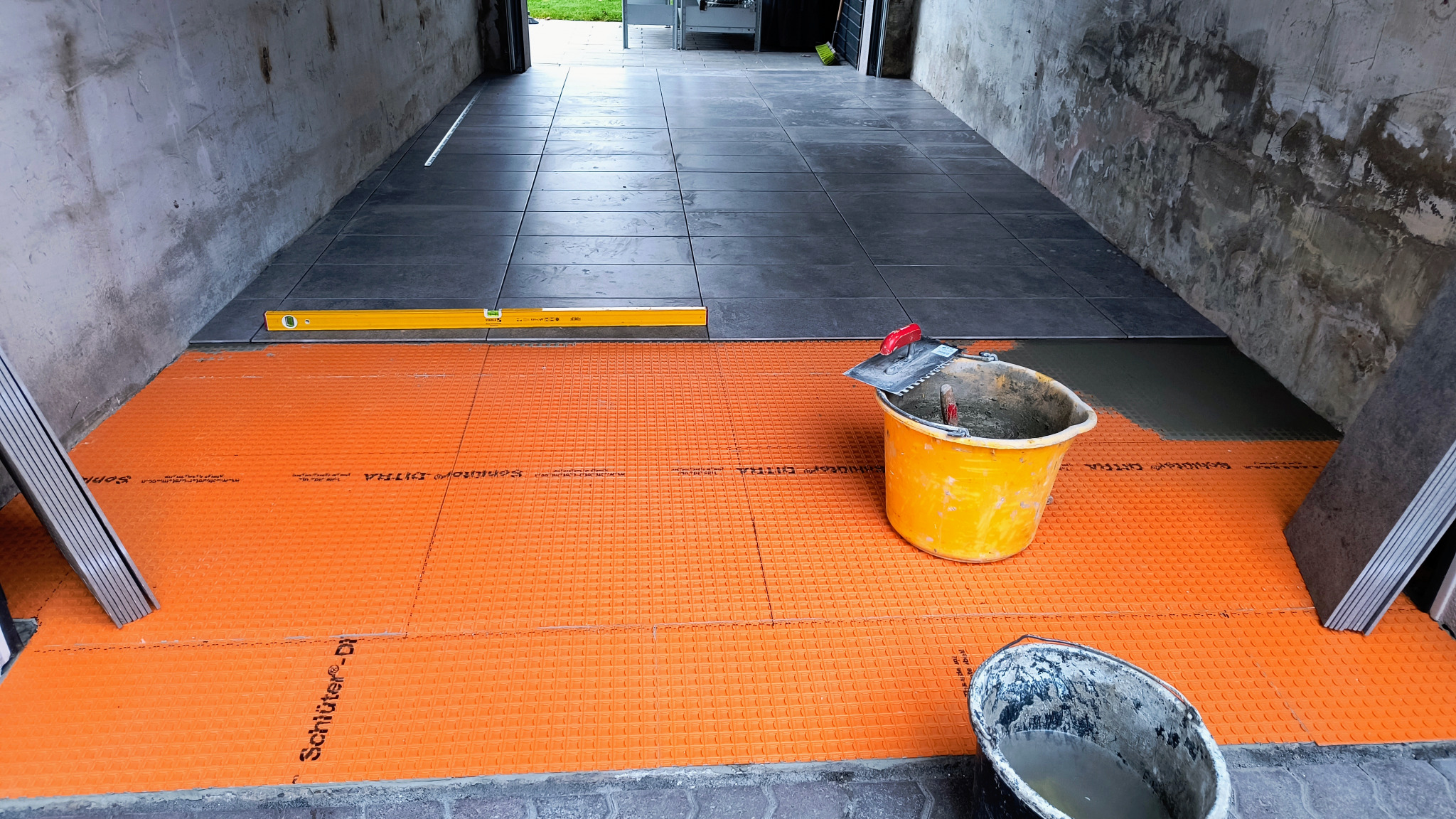 Układanie szarych płytek na pomarańczowej macie drenażowej wewnątrz budynku. Widoczny poziomica, wiadro z zaprawą i płytki. Prace glazurnicze w toku.