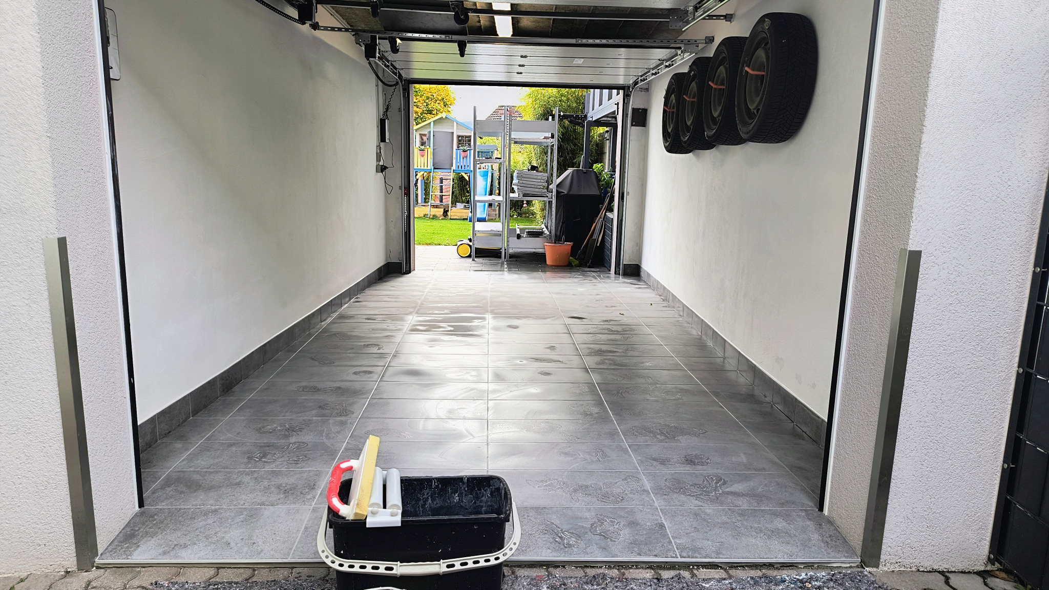 Wykończone wnętrze garażu z szarymi płytkami podłogowymi i białymi ścianami. Widoczny kubeł z narzędziami do czyszczenia płytek i opony samochodowe na ścianie.