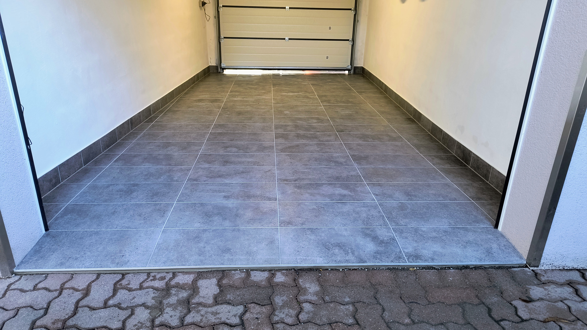 Wykończona podłoga z szarych płytek w garażu z otwartą bramą. Widoczne fugi i listwy przypodłogowe. Świeżo położona podłoga w garażu.