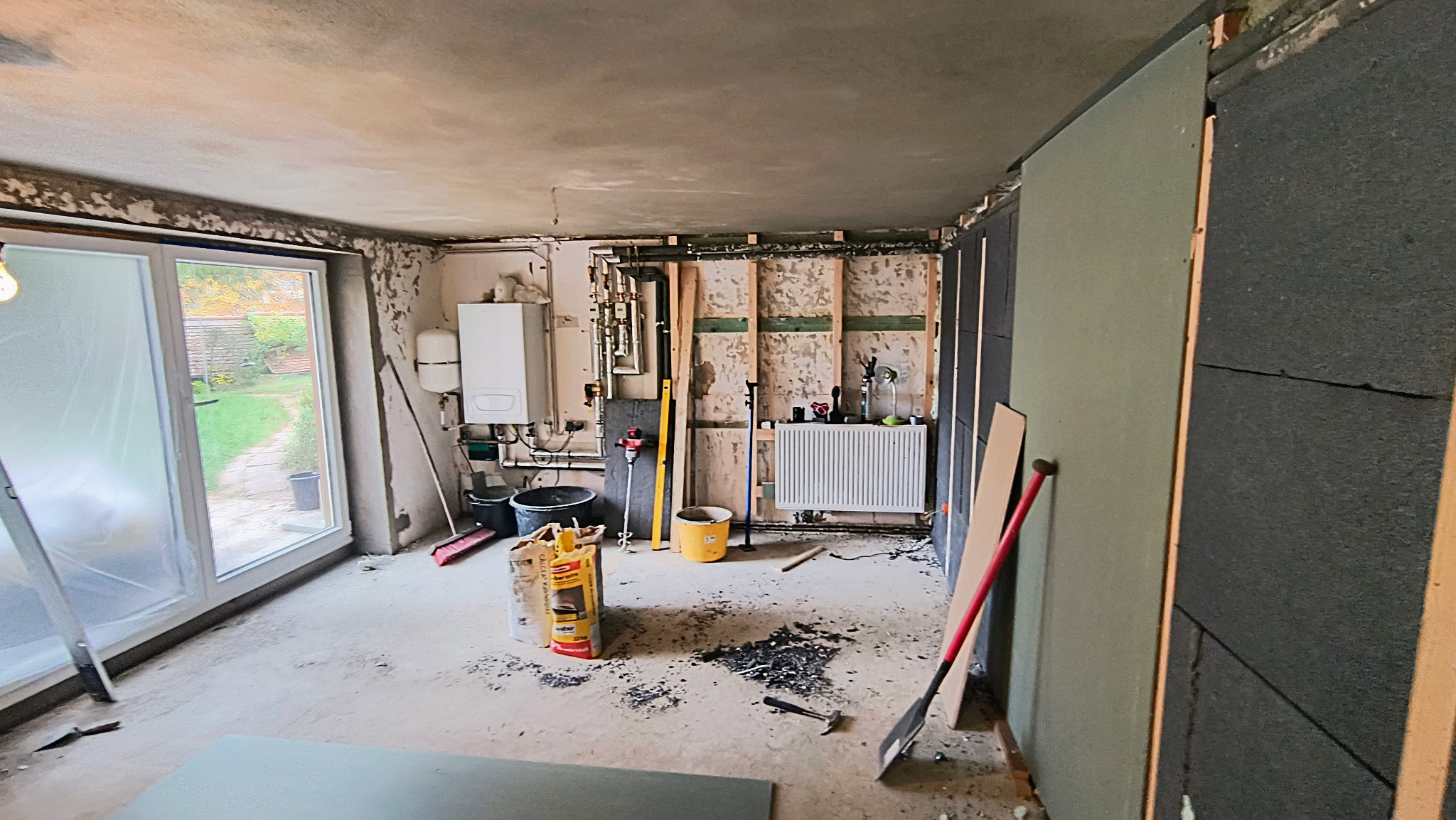 Wnętrze w trakcie remontu: ocieplona ściana z płytami, instalacja grzewcza, okno balkonowe, narzędzia i materiały budowlane. Widoczny etap prac wykończeniowych.