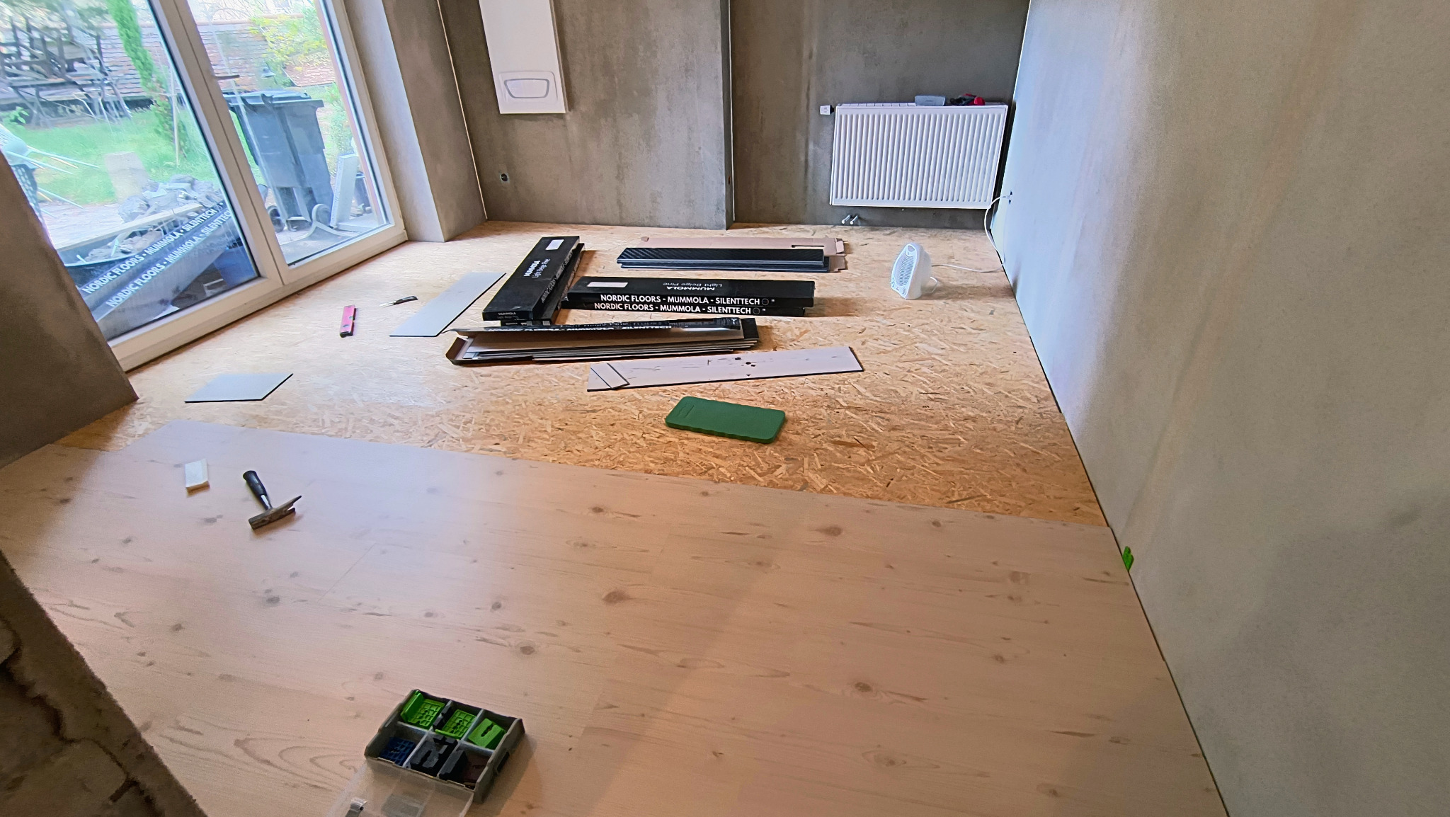 Układanie paneli podłogowych w jasnym wnętrzu. Częściowo ułożona podłoga, widoczne narzędzia, paczki paneli Nordic Floors i grzejnik na ścianie. Etap prac wykończeniowych.