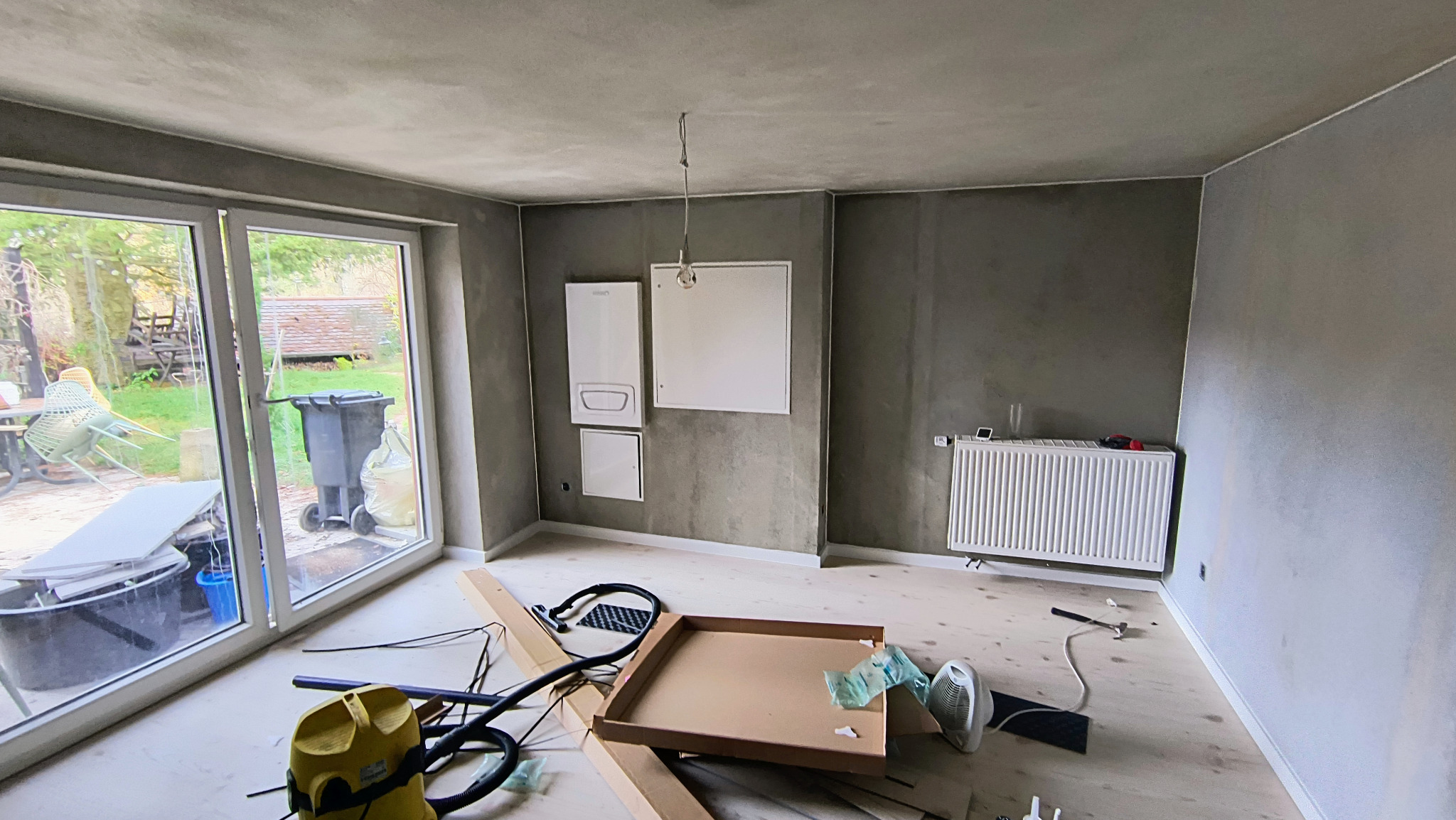 Pokój po tynkowaniu z widocznym grzejnikiem, oknem balkonowym i narzędziami. Ściany w odcieniach szarości, surowy wygląd. Widok na zewnątrz przez okno.
